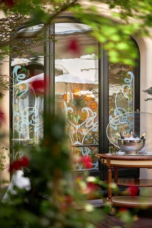 A room with a glass door featuring floral designs, a table with a metal bowl containing bottles, and a view of a garden through the door. - Hotel Regina Louvre (Paris)