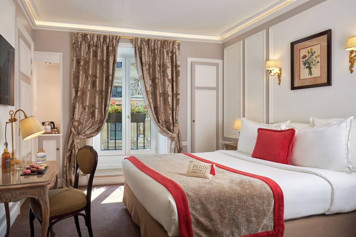 A hotel room with a bed, a desk with a lamp, a chair, and a window with curtains.