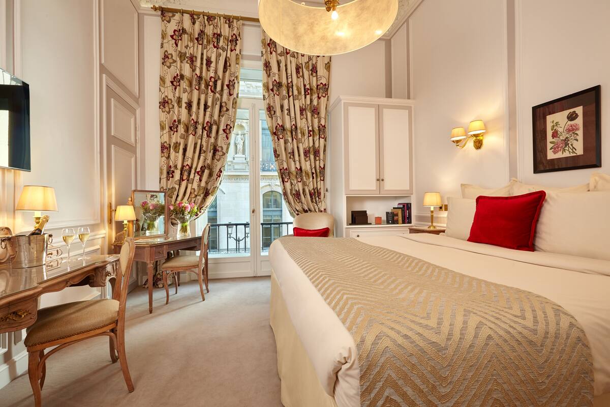 A hotel room with a large bed, a desk with a chair, a dresser, and a view of the outside through the curtains.