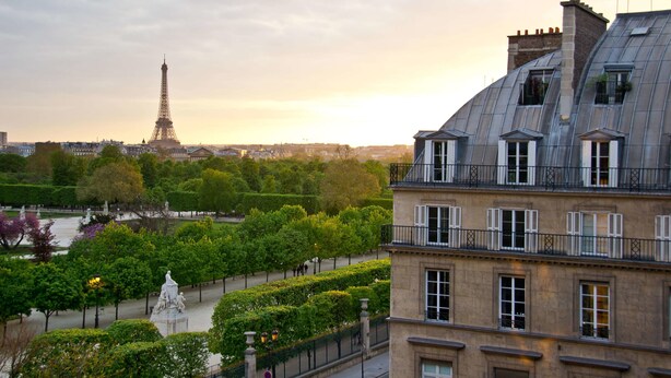 Vista de la Torre Eiffel desde un edificio de techo inclinado, una estatua al frente y un parque con árboles y una fuente.