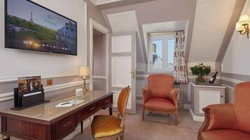Camera d'albergo con una grande TV a schermo piatto che mostra una scena parigina, una scrivania in legno con una lampada, due poltrone imbottite e un tavolino con un vaso e una bottiglia.