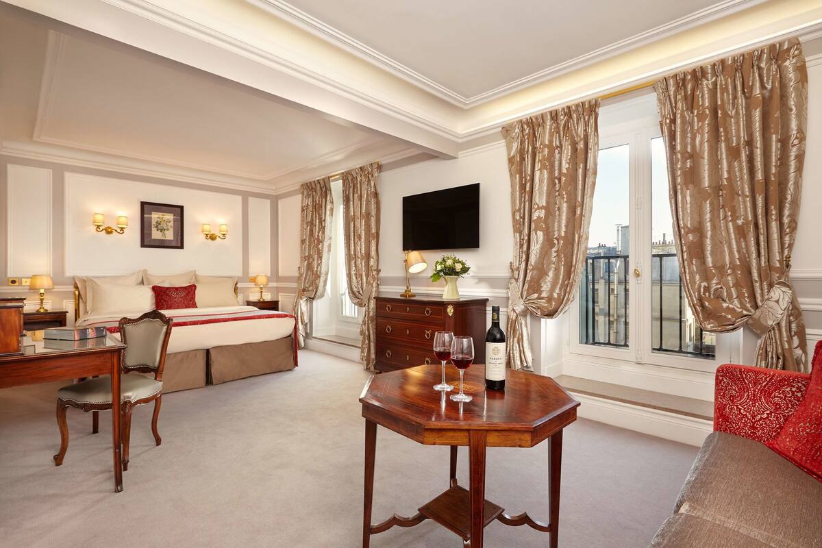 A hotel room with a bed, a desk with a chair, a TV mounted on the wall, and a window with curtains.
