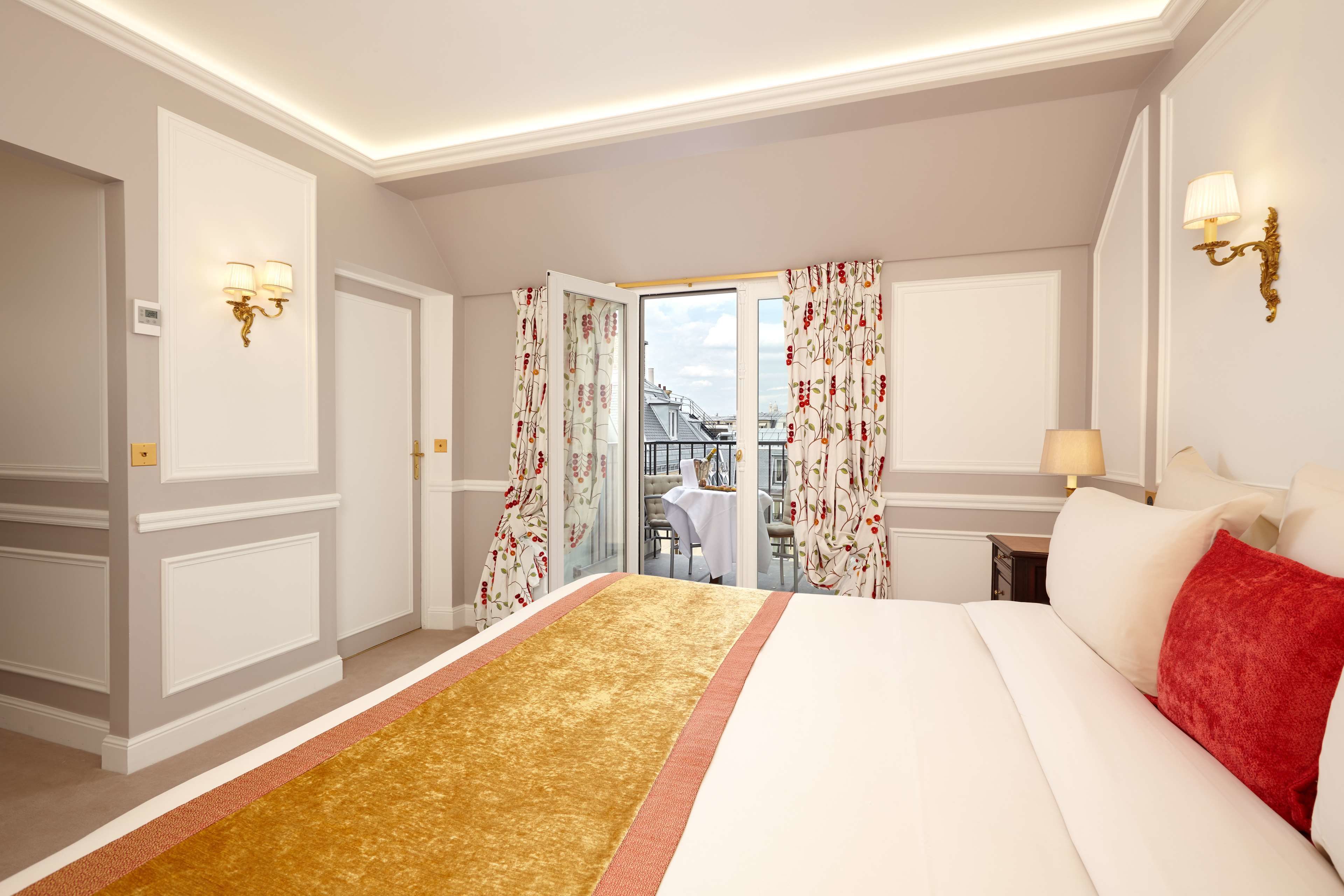 Ein Hotelzimmer mit einem großen Bett, einem Teppich mit goldenen Akzenten, Blick durch eine geöffnete Tür und einem Balkon mit Tisch und Stühlen.