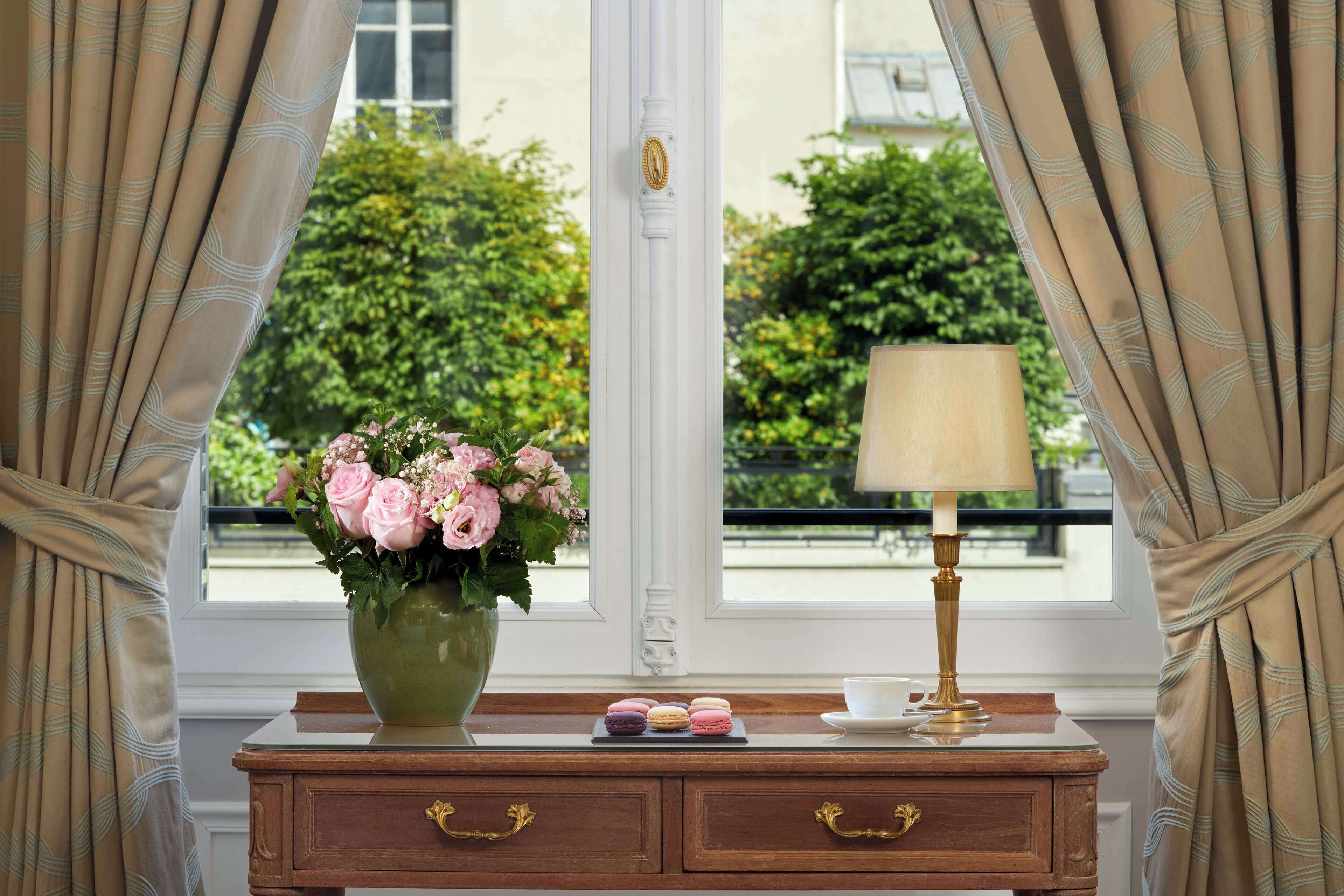 A wooden desk with a vase of pink flowers, a lamp, and macarons.