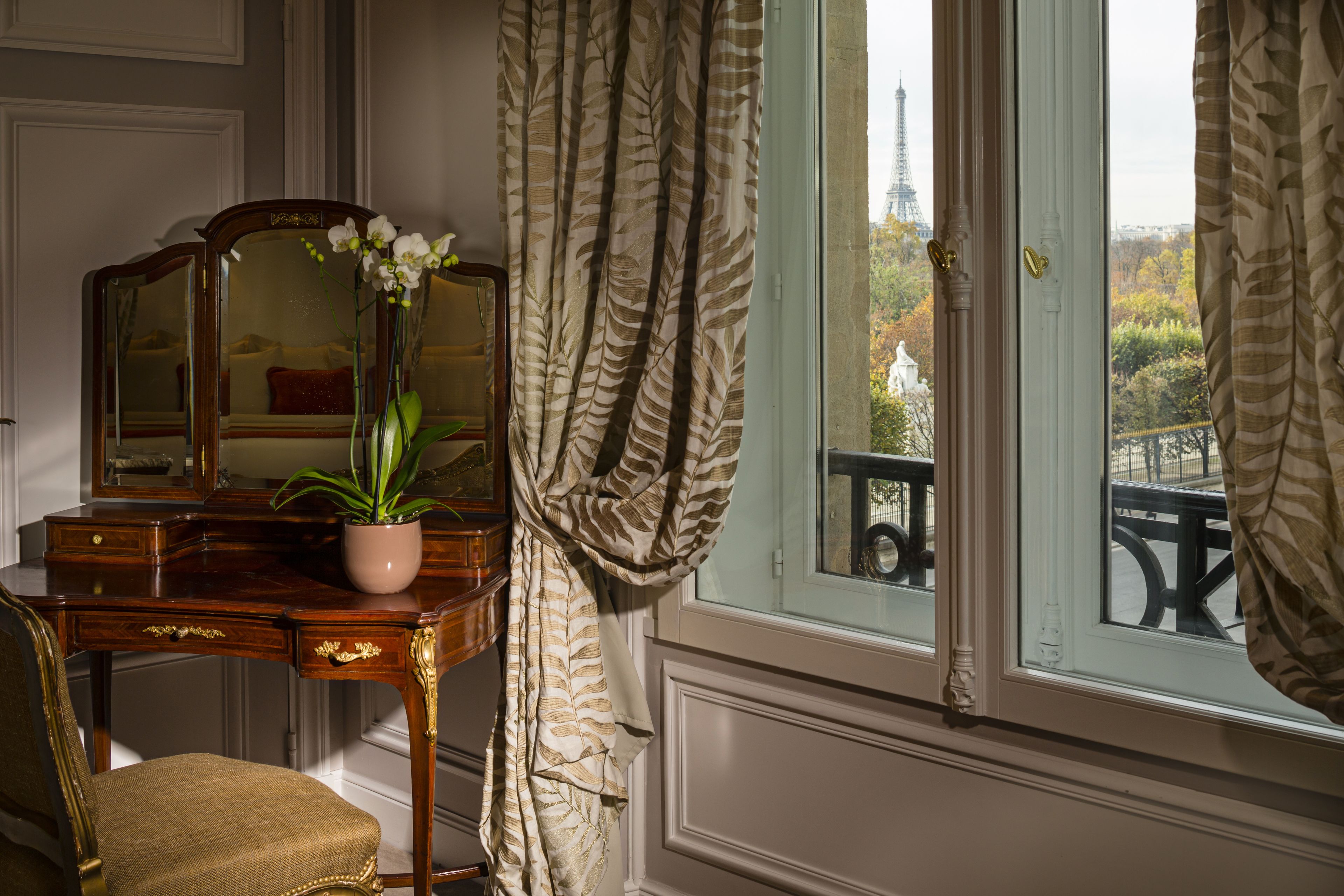 Une pièce avec un bureau en bois, une chaise, un miroir et une vue sur la Tour Eiffel, visible à travers une fenêtre ornée de rideaux.