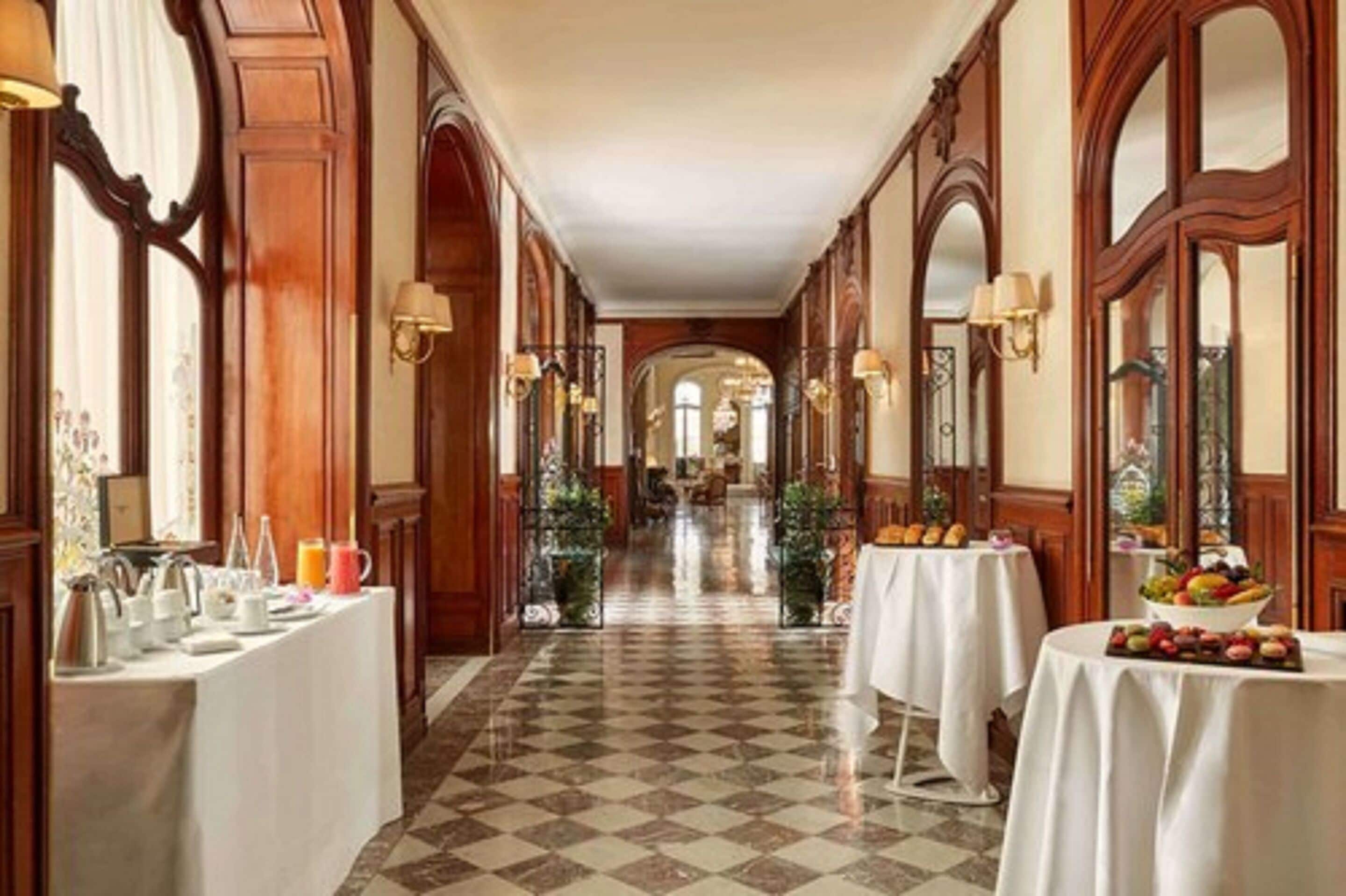 A hotel corridor with checkered flooring, wooden paneling, and tables set with breakfast items.