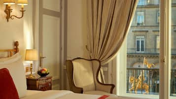 Habitación de hotel con cama, mesita de noche con lámpara, una silla y vistas a un edificio con balcones.