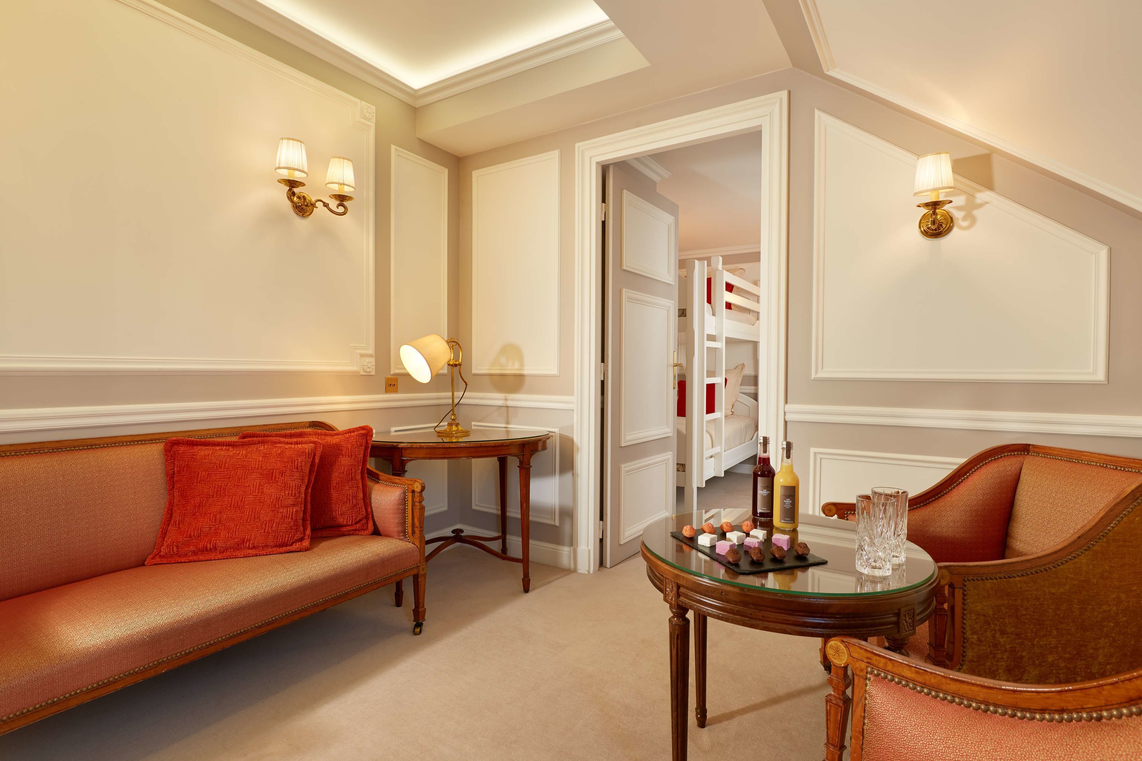A hotel room with a leather sofa, a small round table with a lamp, and a glass-top table with bottles and glasses.
