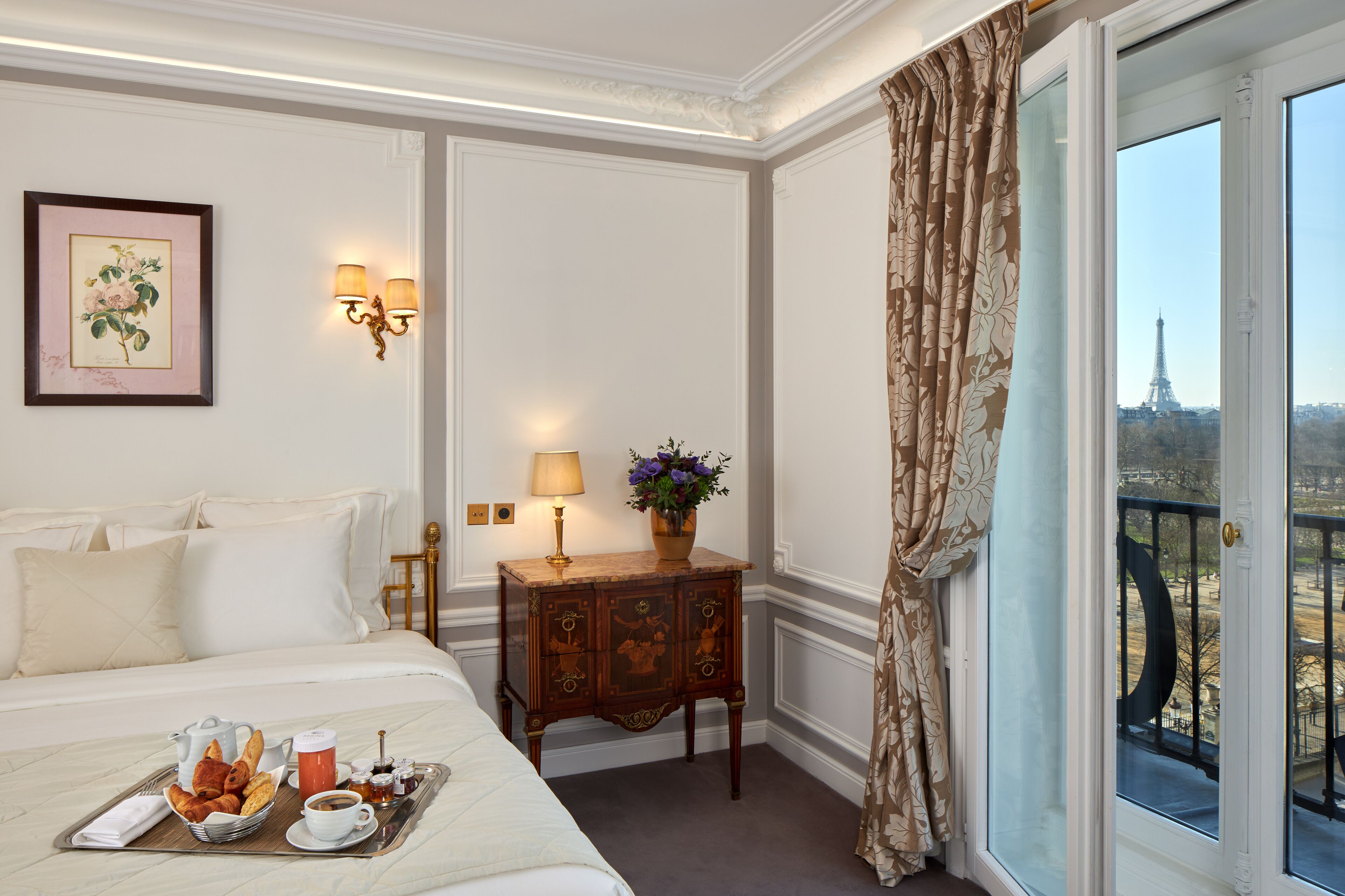 A hotel room with a bed, breakfast tray, a small table with a lamp, a decorative cabinet, and a view of the Eiffel Tower.