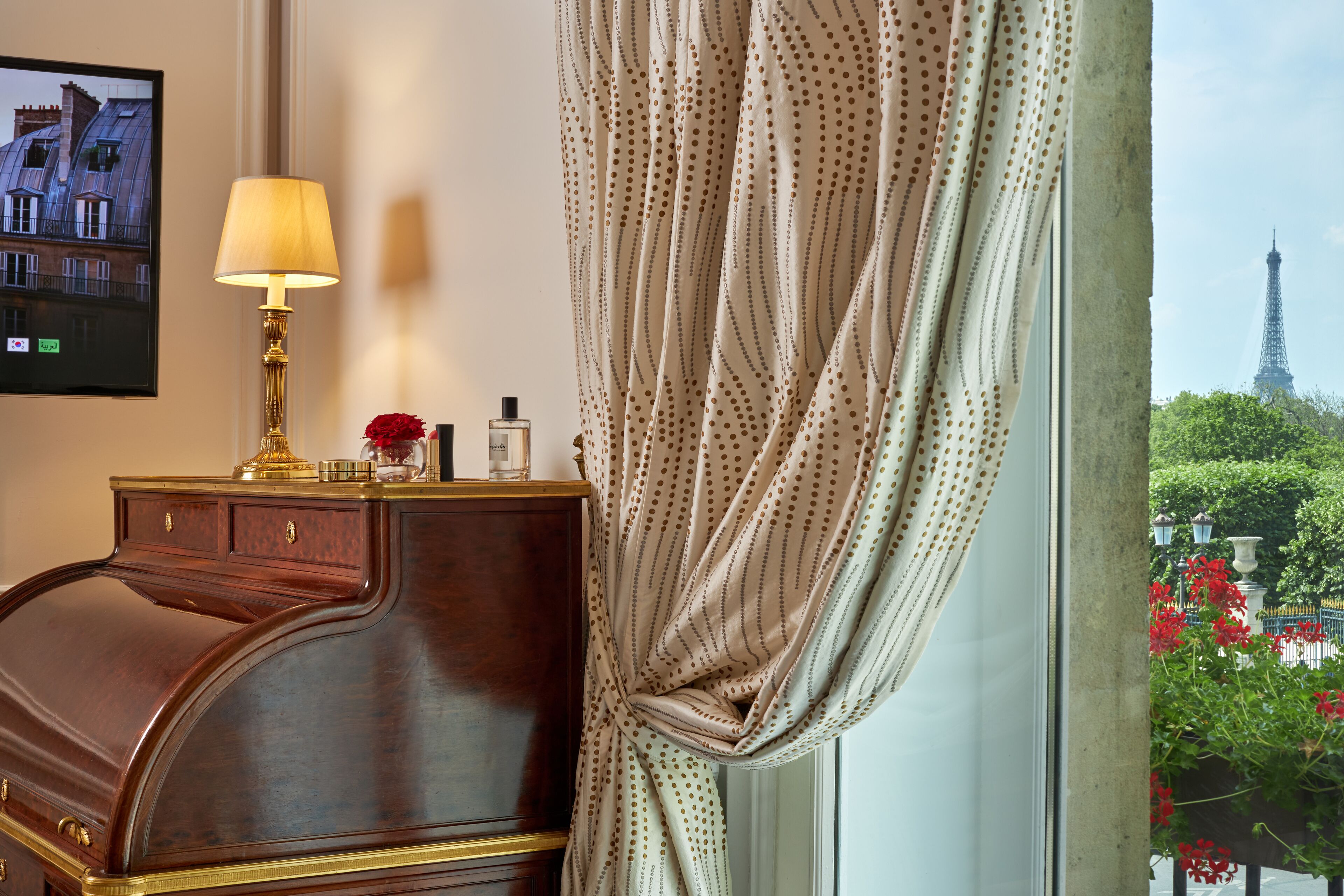 A room with a wooden desk, a lamp, and a view of the Eiffel Tower through a window with curtains.