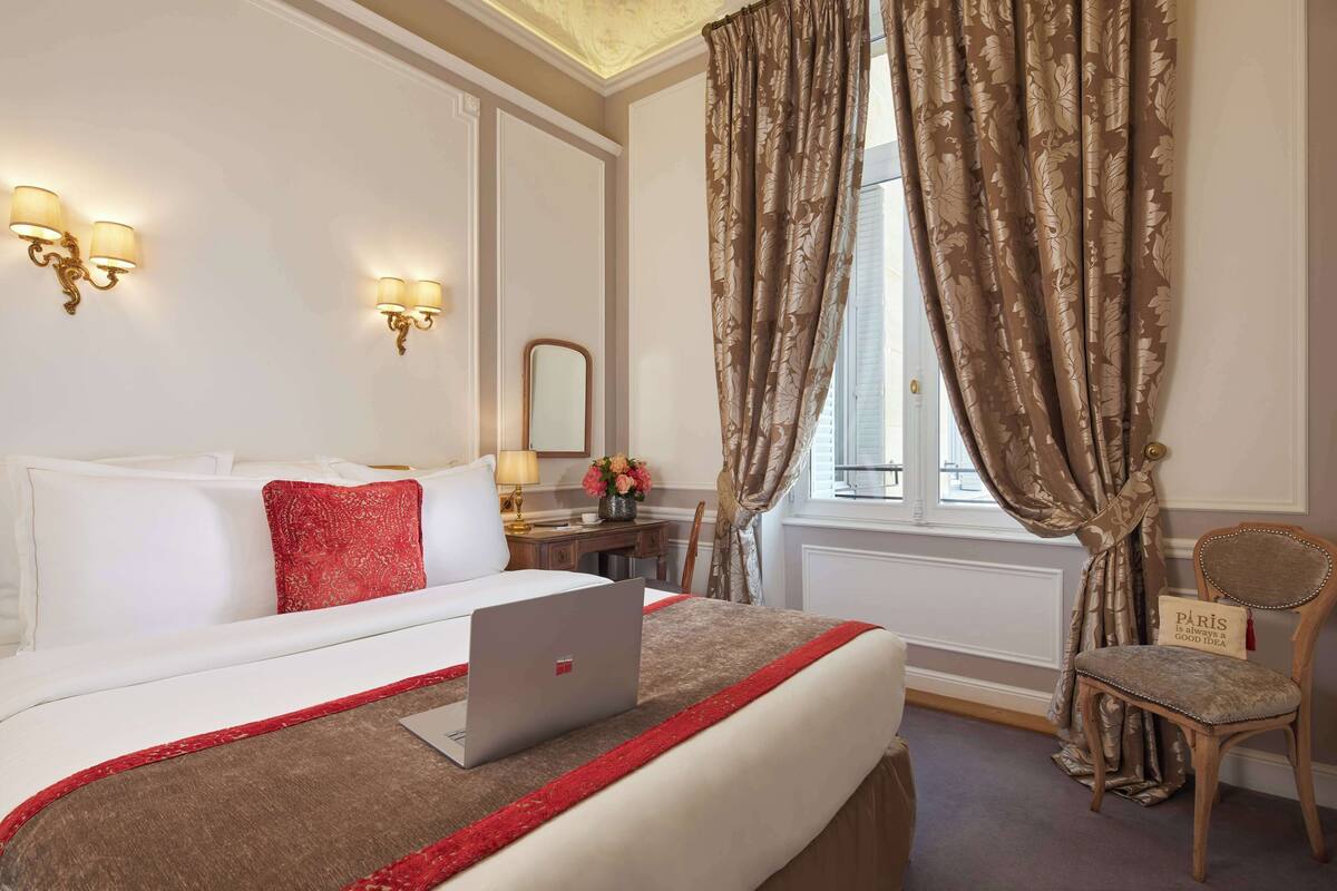 A hotel room with a bed, a laptop, a chair, a desk, and a window with curtains.
