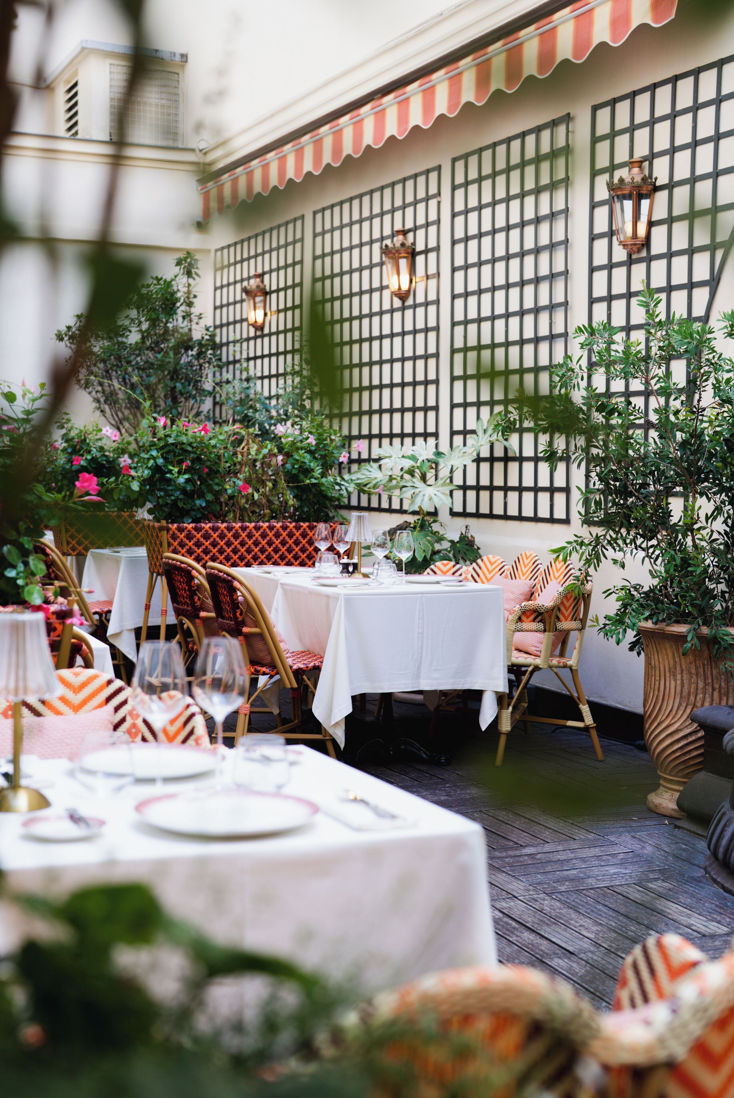 An outdoor dining area with tables set for two, surrounded by greenery and potted plants.