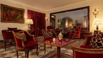 A luxurious room with a checkered floor, red upholstered chairs, a wooden table, and a large mirror reflecting a garden view.