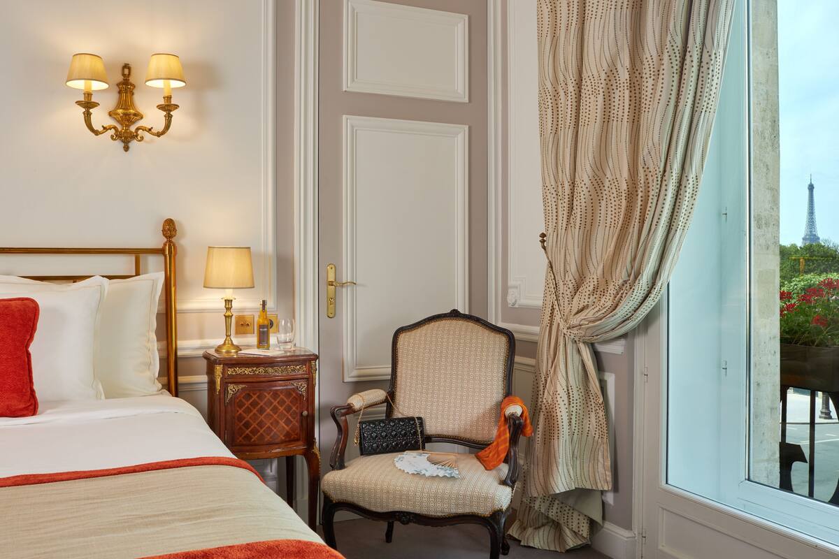 A hotel room with a bed, a chair, a nightstand, and a view of the Eiffel Tower.