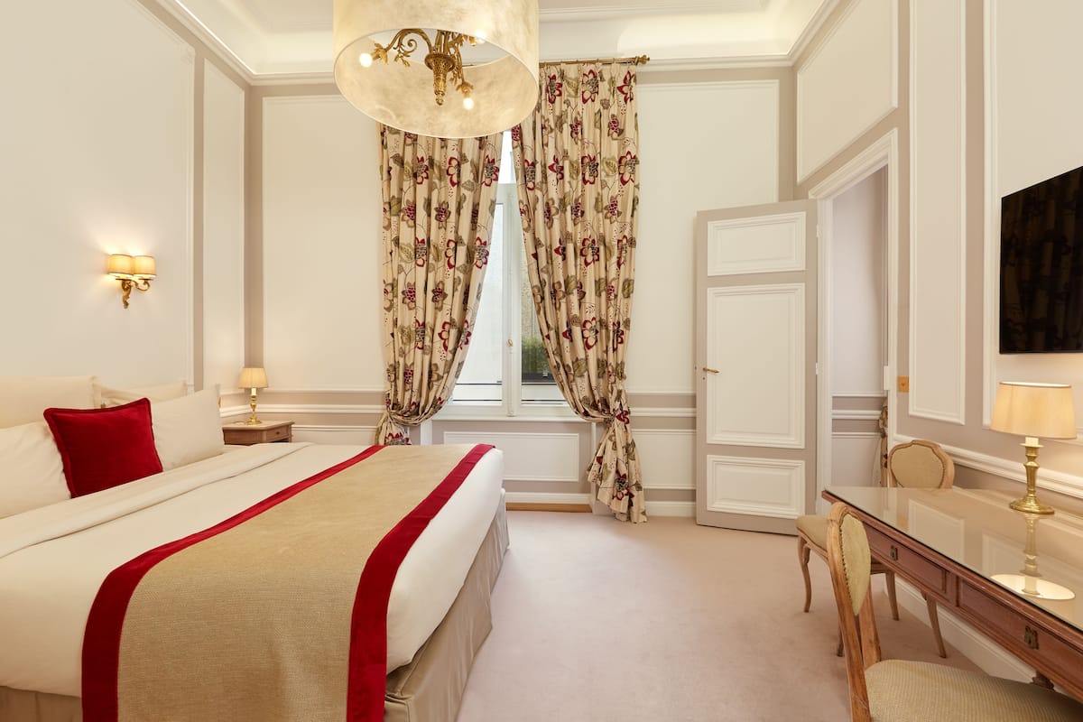 A hotel room with a large bed, a desk, and a chair. There is a window with curtains, a lamp on the desk, and a painting on the wall.