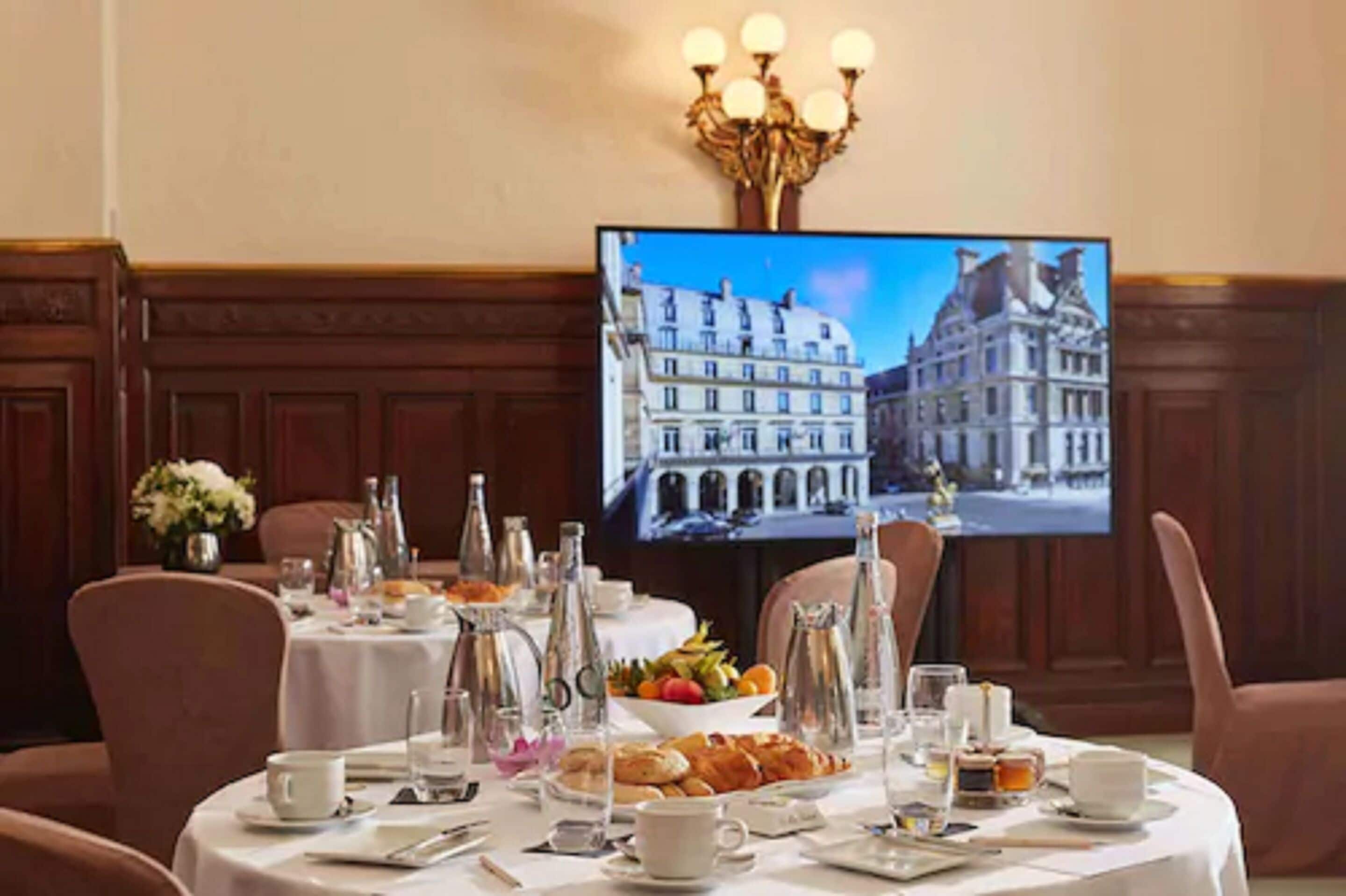Un comedor con mesas redondas dispuestas para el desayuno, una pantalla de televisión mostrando un paisaje urbano y una araña colgante.