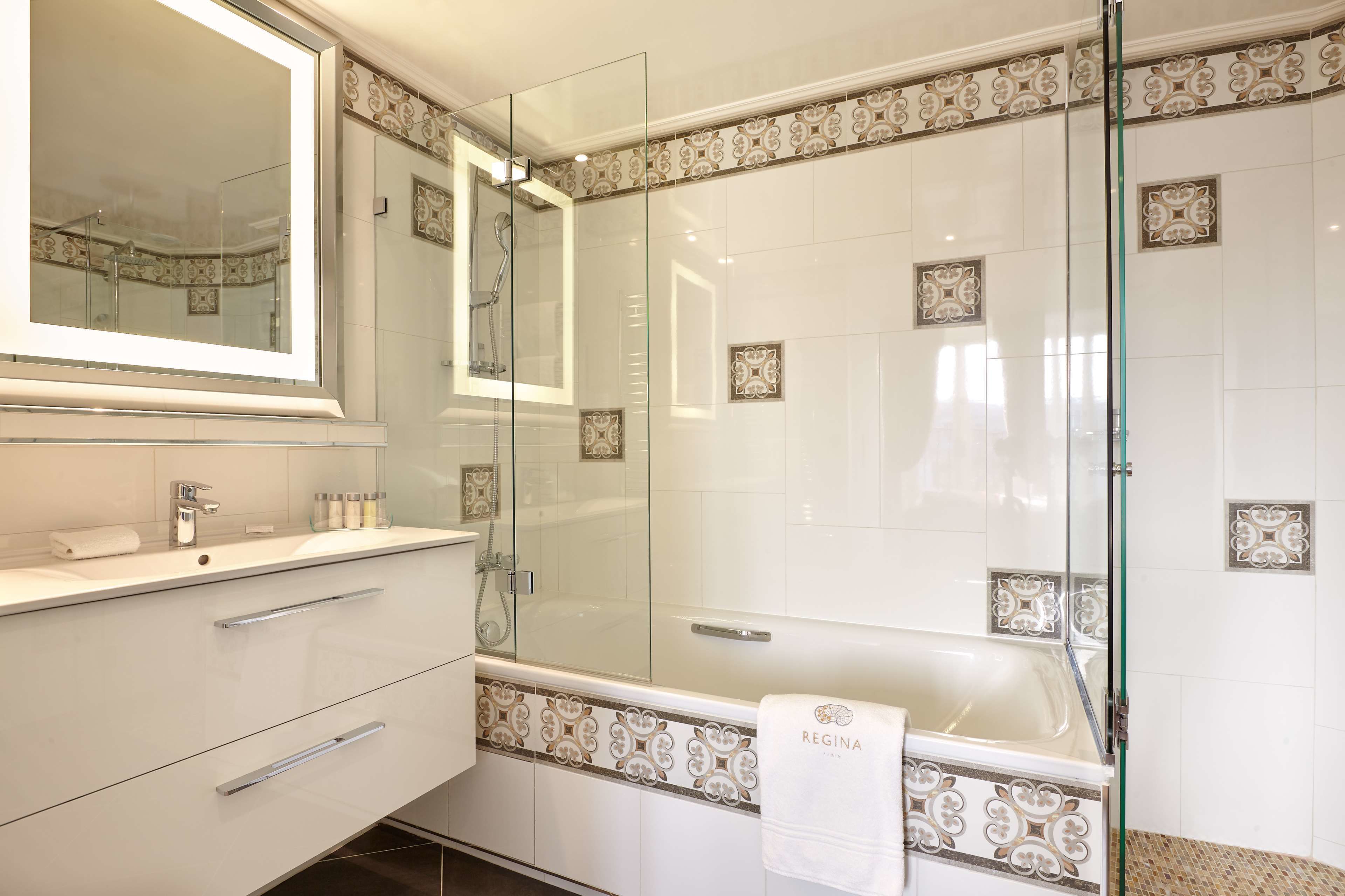 A modern bathroom with a glass-enclosed shower, a large mirror, and a bathtub with a decorative tile border.