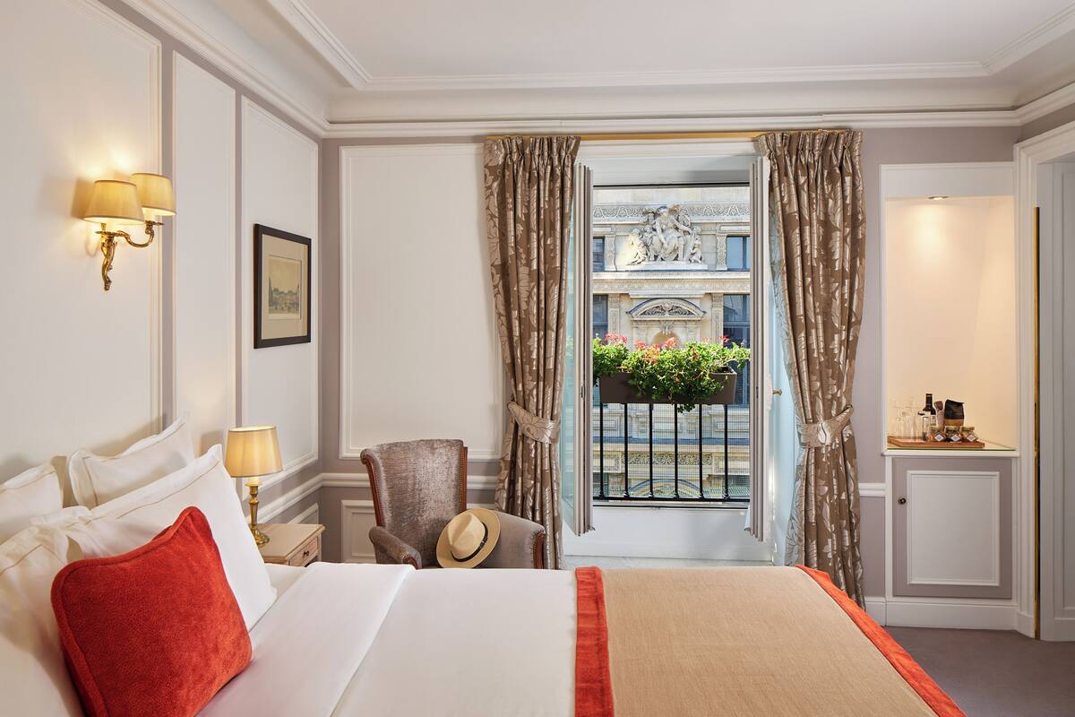A hotel room with a bed, a chair, a nightstand, a lamp, a window with curtains, and a view of a balcony with plants.