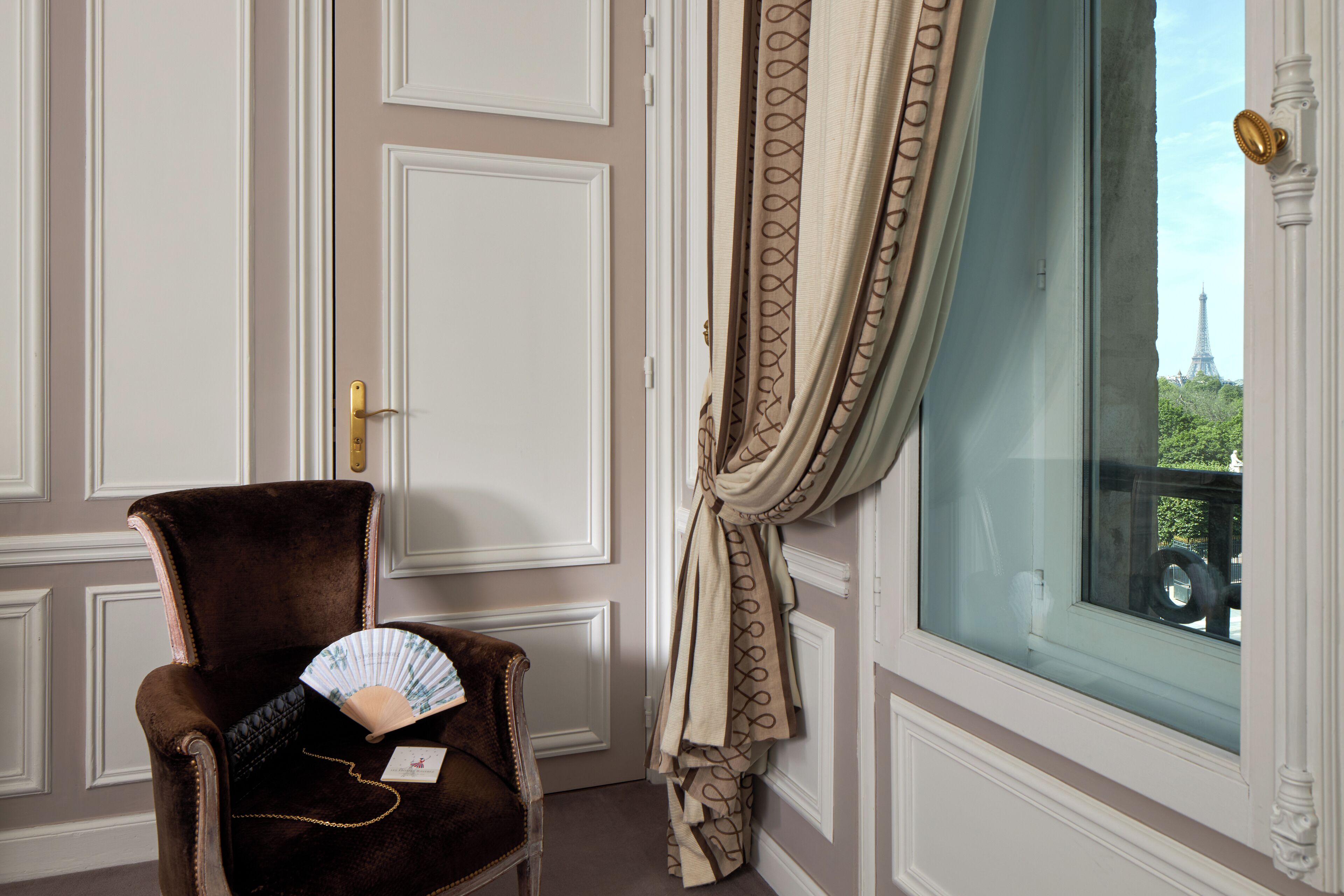 A room with a brown armchair, a window with a view of the Eiffel Tower, and a door with a gold handle.