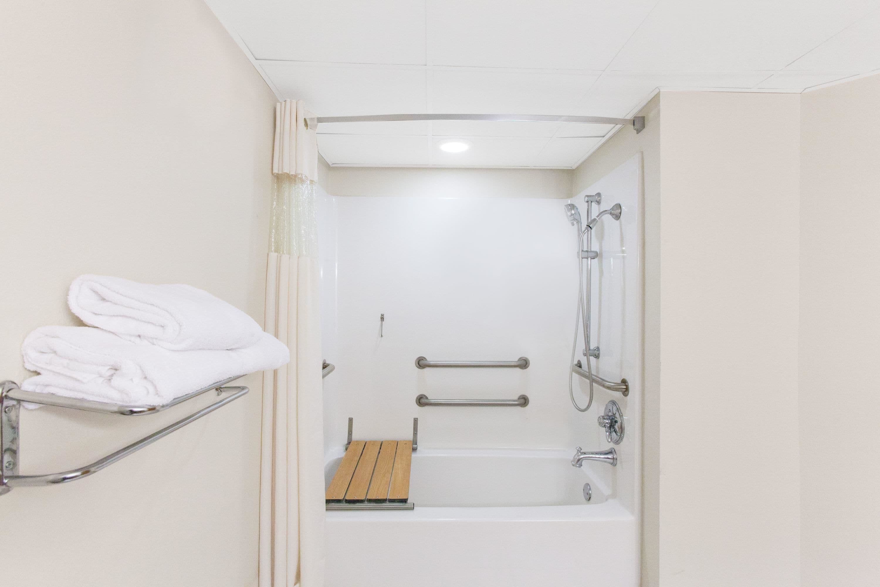 A small, white bathroom with a shower, a wooden shelf with towels, and a grab bar.