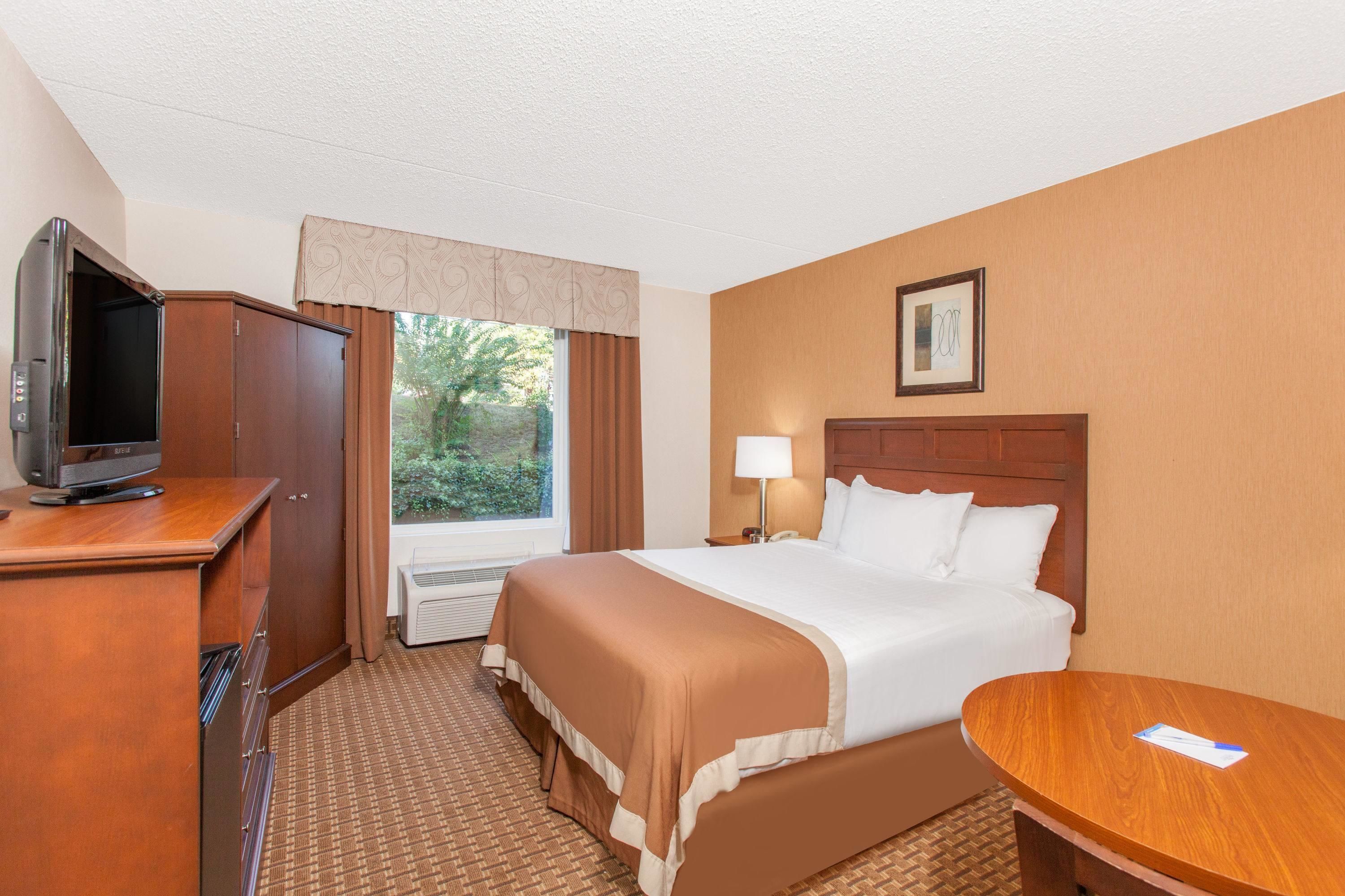 Habitación de hotel con una cama, un escritorio con televisor, una lámpara y una ventana con cortinas.