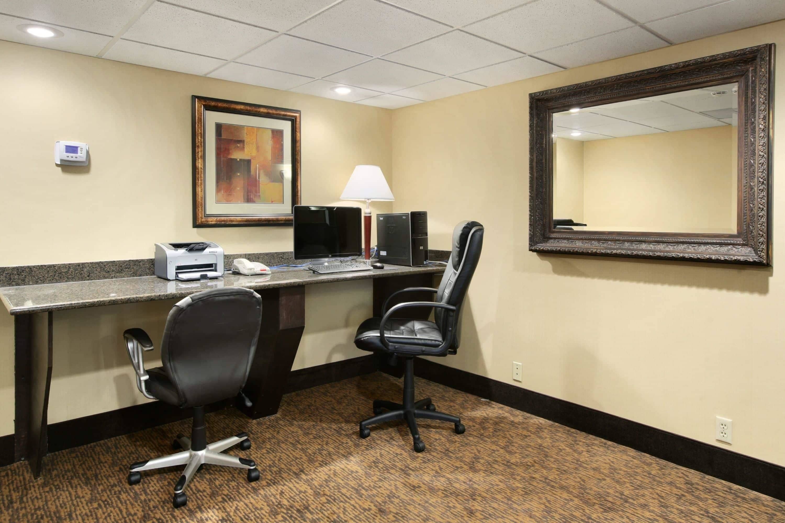 A hotel room with a desk, two office chairs, a computer, a printer, a lamp, and a large mirror.