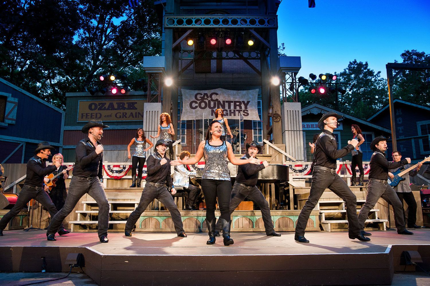 Un escenario con un grupo actuando, con un cartel que dice "GAC COUNTRY NIGHTS" y un edificio al fondo.