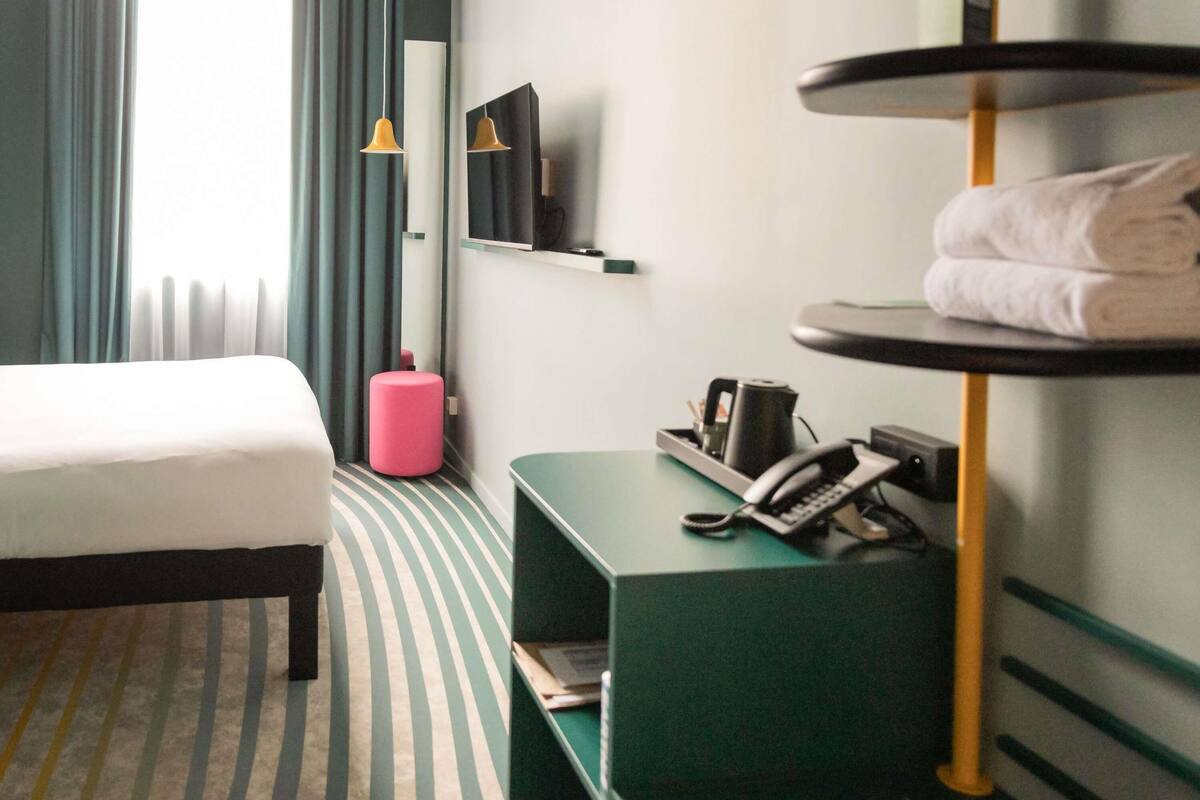 A hotel room with a bed, a small desk with a telephone, a pink stool, and a wall-mounted TV.
