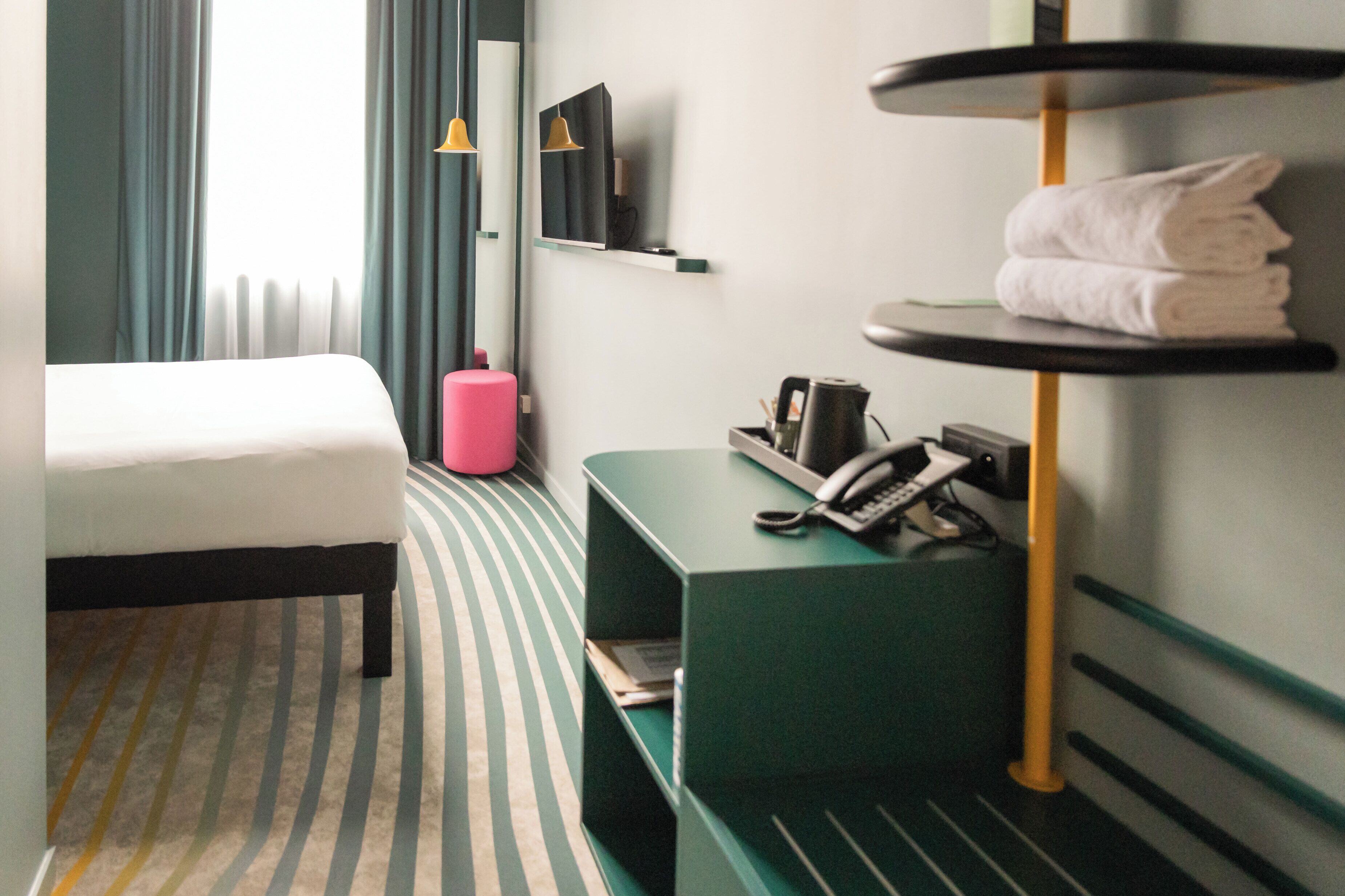 Een hotelkamer met een bed, een groen nachtkastje met een telefoon, een roze kruk en een wandplank met handdoeken.