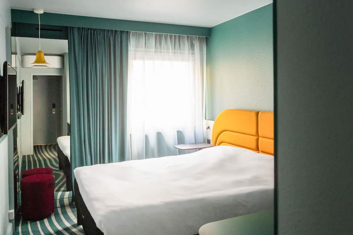 Quarto de hotel com uma cama grande, uma mesa pequena, uma cadeira, um banquinho vermelho e uma janela com cortinas.