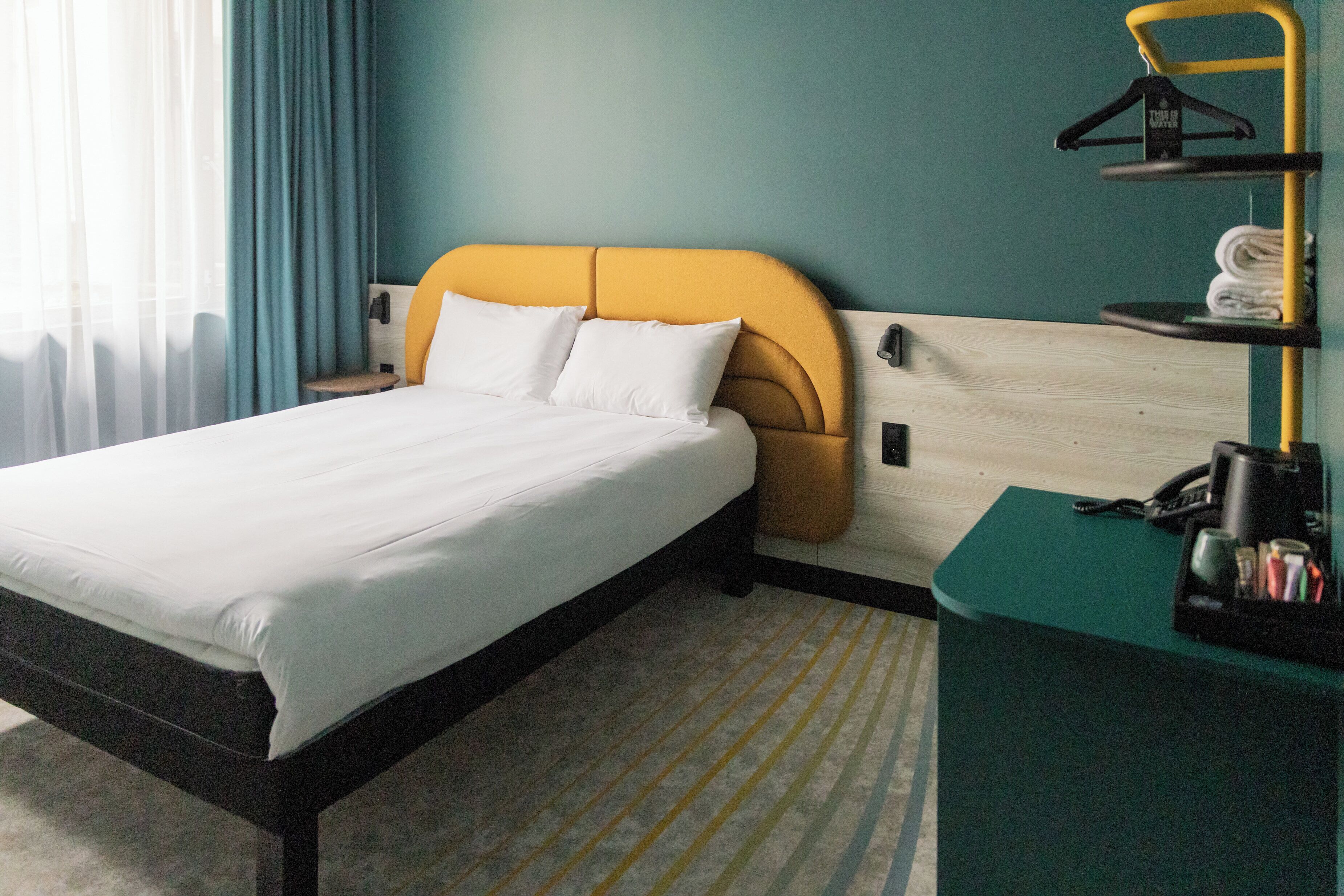 Habitación de hotel con cama, cabecera de madera, escritorio con teléfono y estante con toallas.