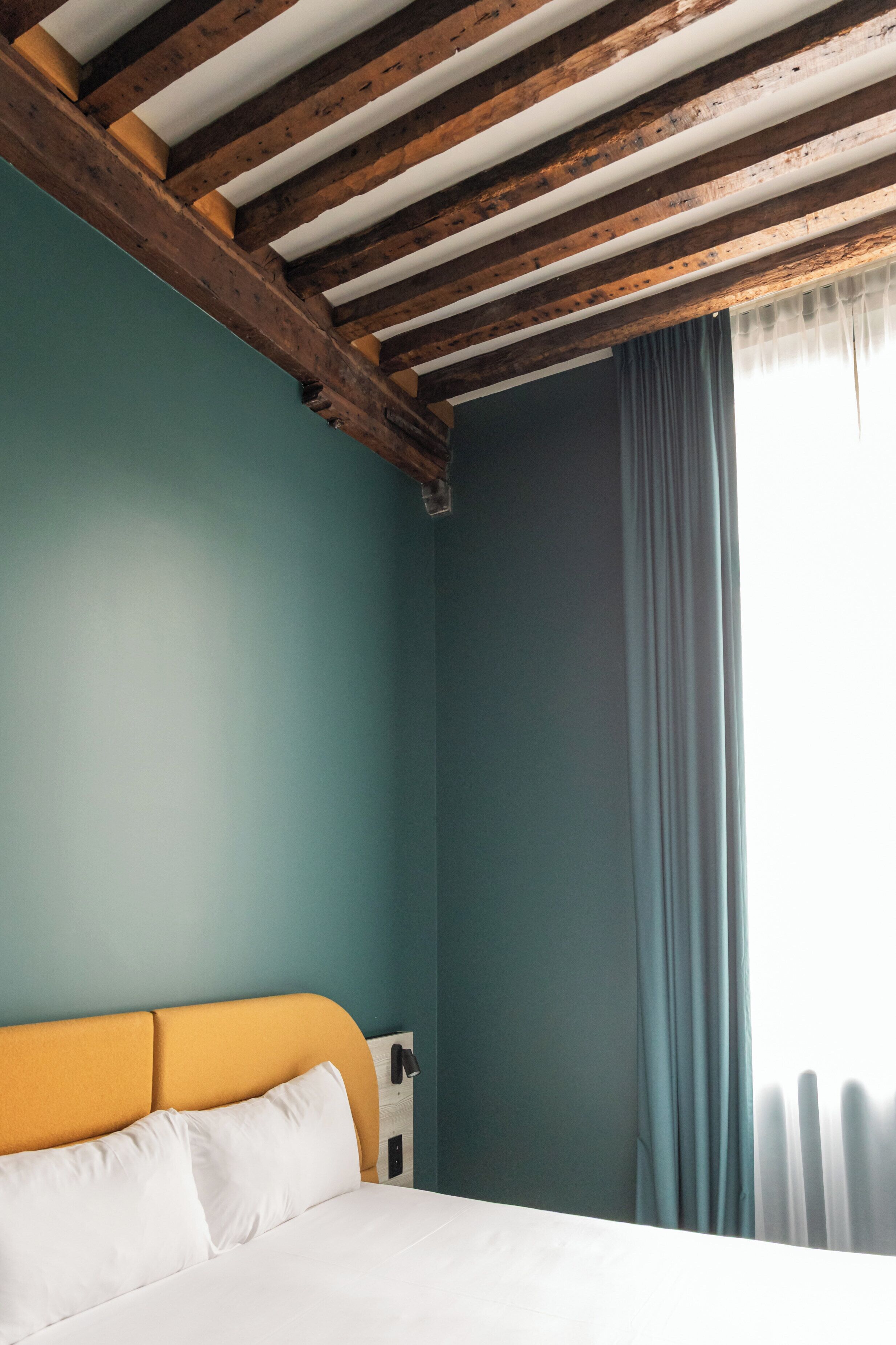 Un dormitorio con techo de madera, pared verde azulado, cabecera amarilla y cama blanca.