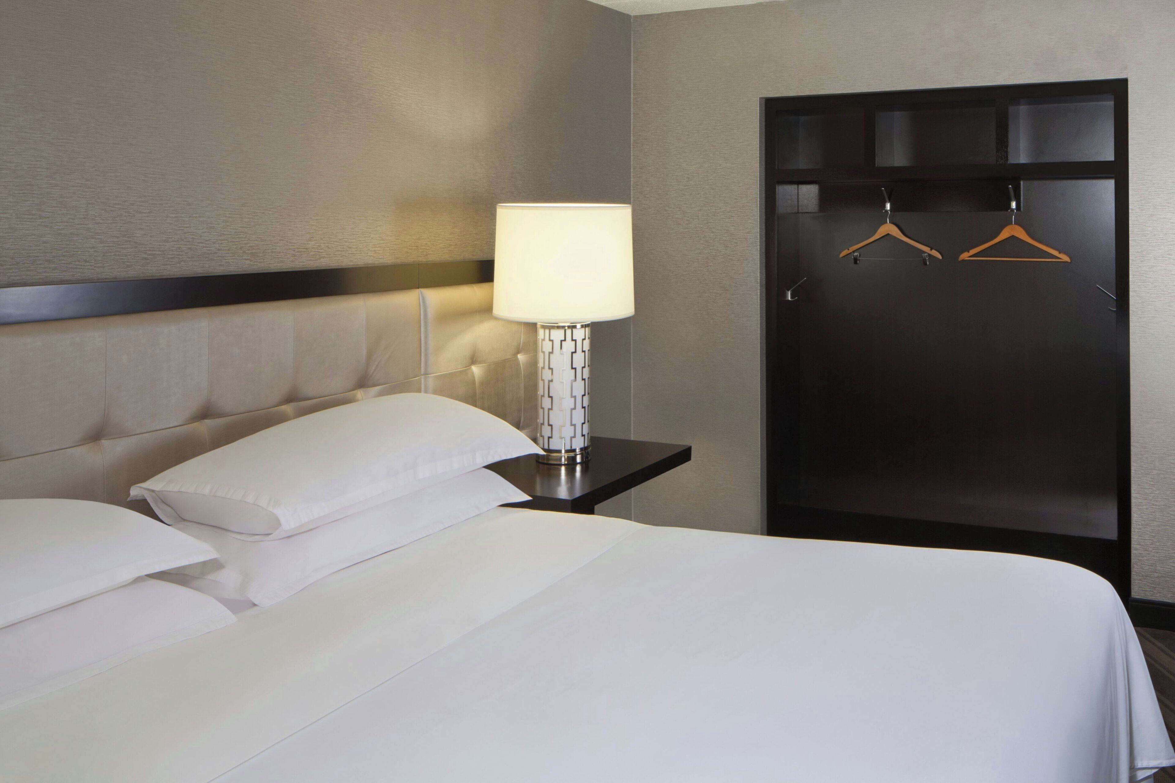 Habitación de hotel con cama, almohadas, mesita de noche con lámpara y armario con perchas.