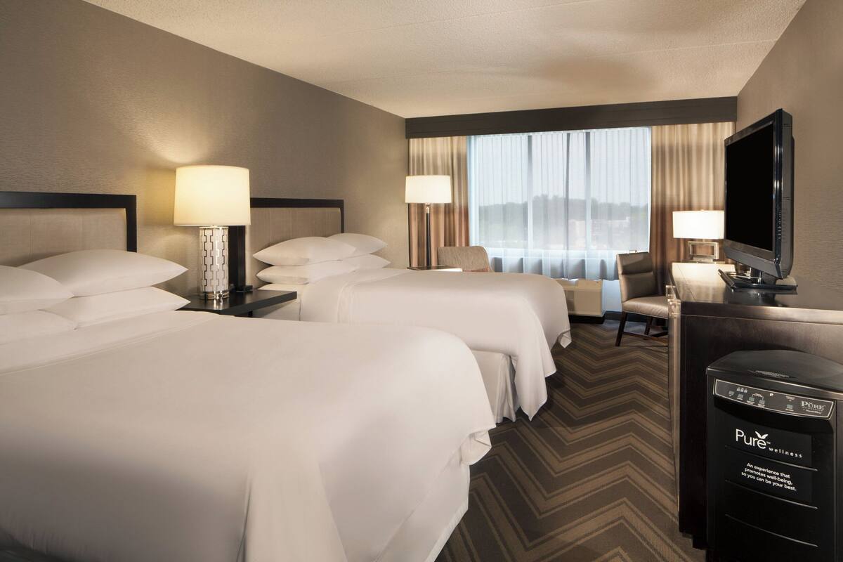 A hotel room with two beds, a desk with a television, and a view of the city through large windows.