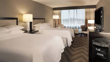 Habitación de hotel con dos camas, un escritorio con televisor y vistas a la ciudad a través de amplias ventanas.