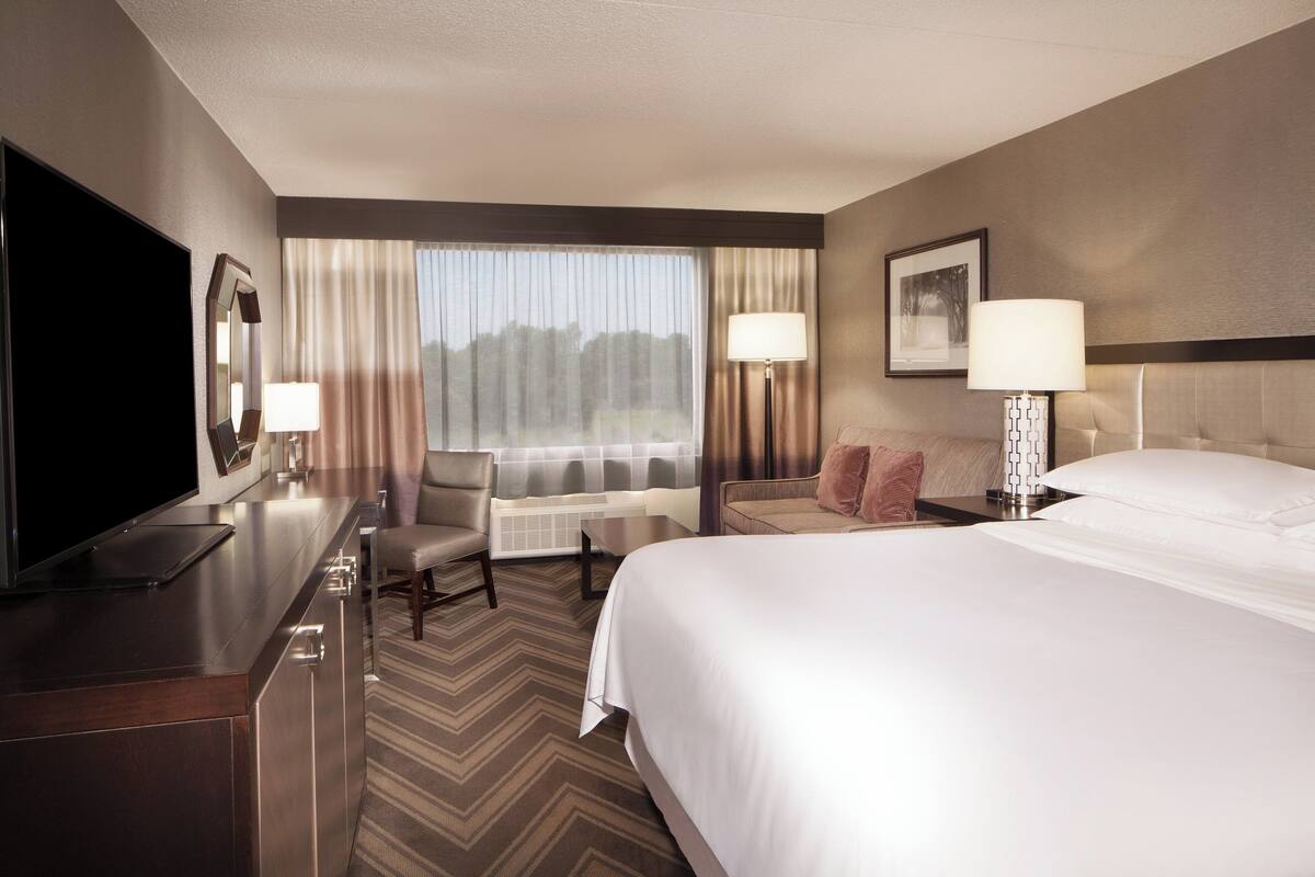 A hotel room with a large bed, a desk with a flat-screen TV, a chair, a sofa, and two lamps.
