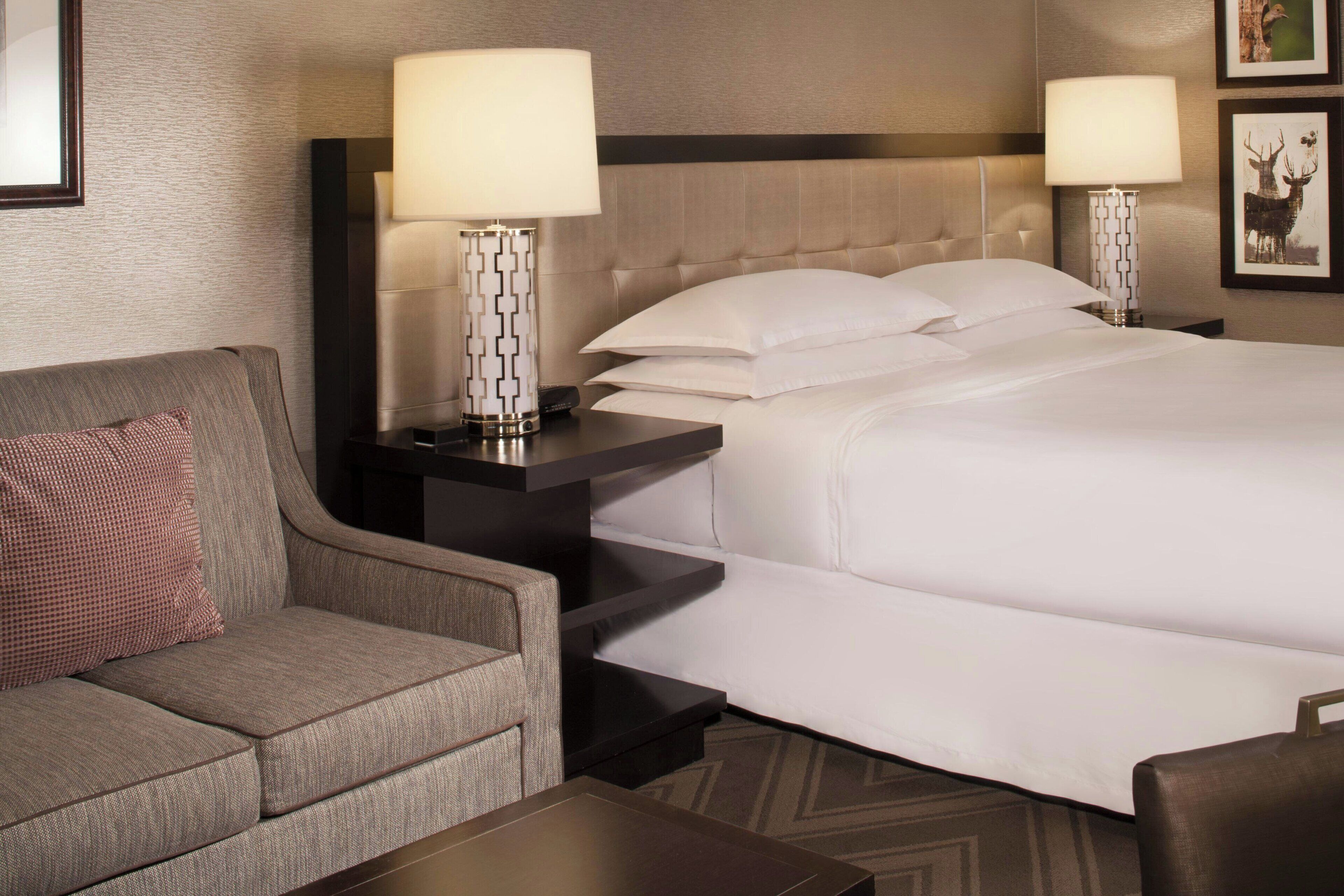 A hotel room with a bed, a sofa, two bedside tables with lamps, and framed pictures on the wall.