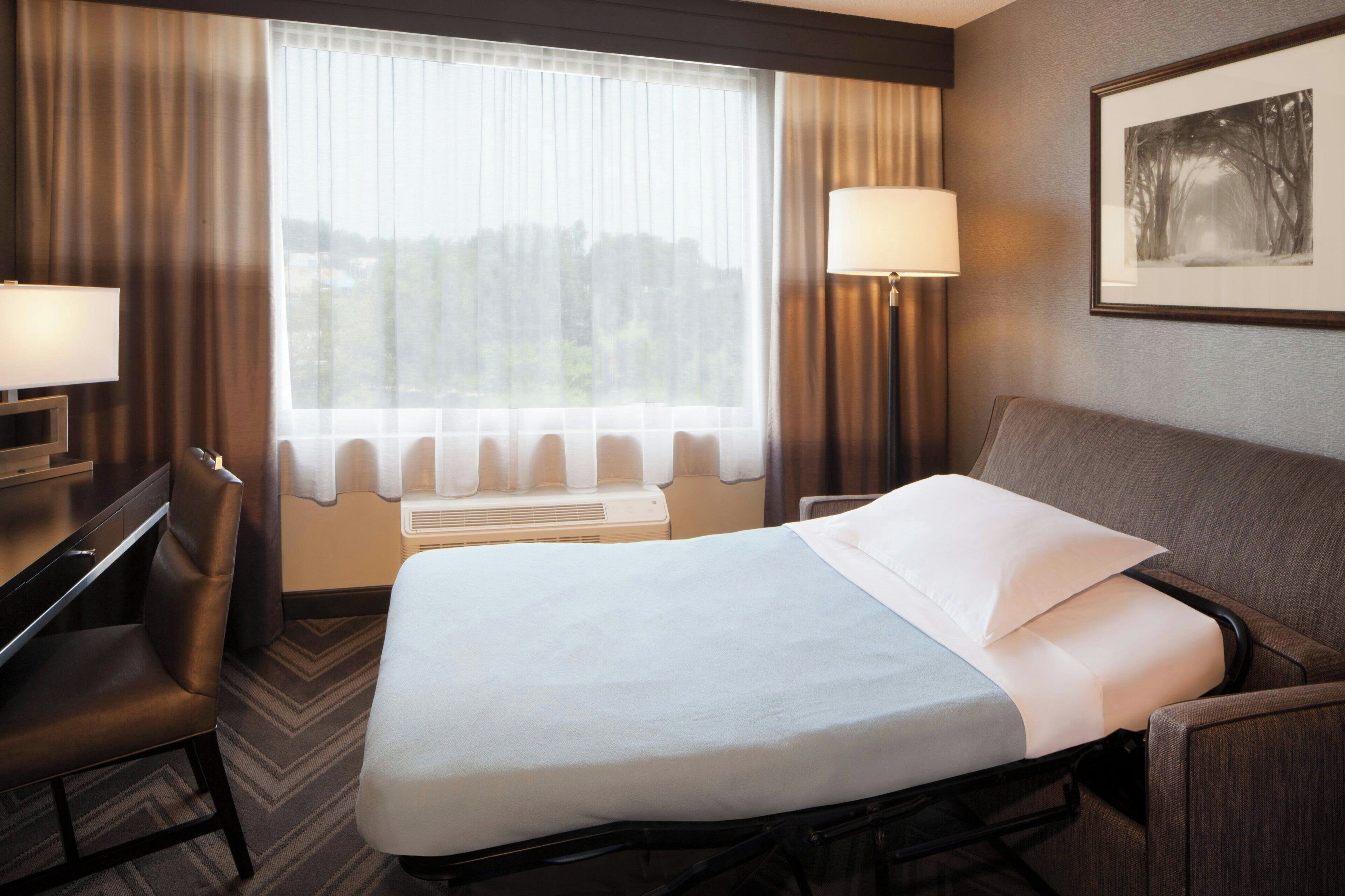 A hotel room with a bed, a desk, a chair, a window with curtains, a wall-mounted lamp, and a framed picture.