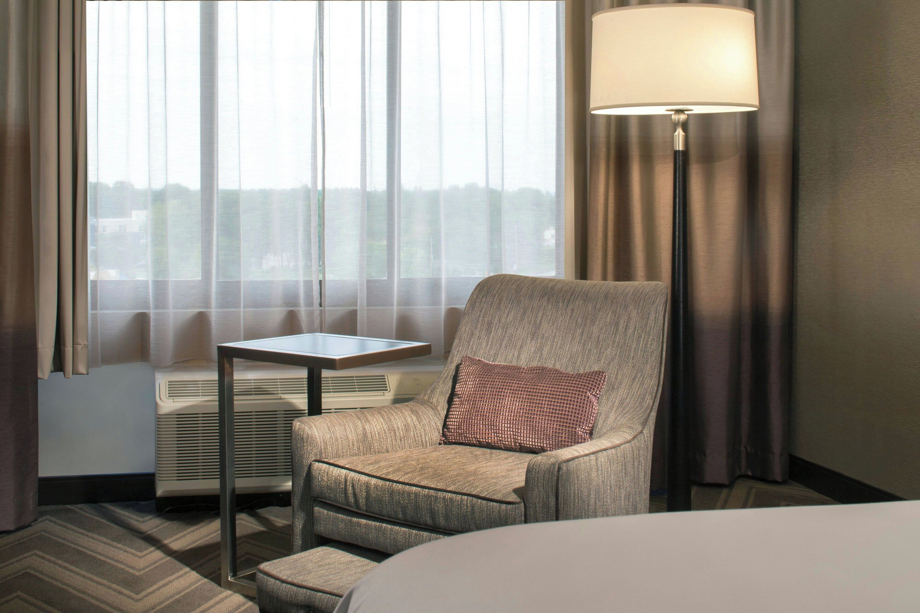 A hotel room with a large window, a chair with a pillow, a small table, a lamp, and a bed.