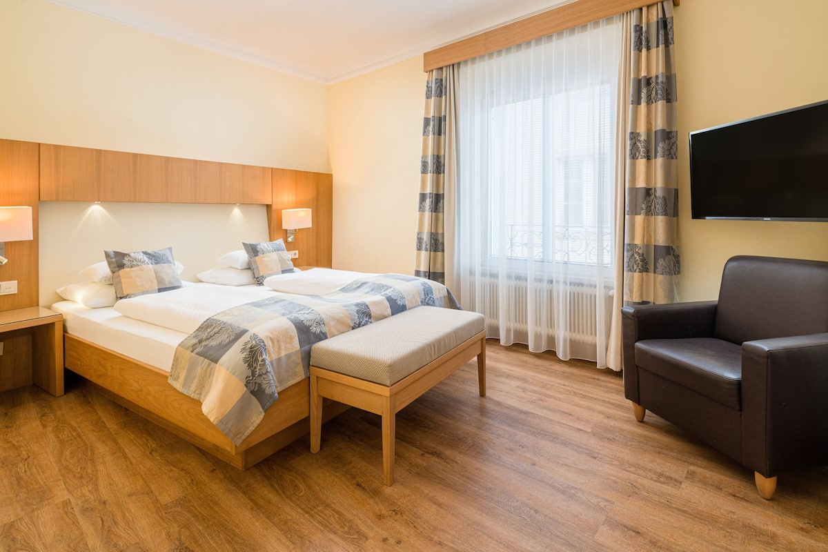 Habitación de hotel con una cama grande, mesitas de noche, una silla, un televisor y una ventana con cortinas.