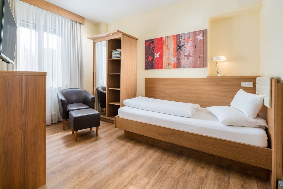 Una habitación de hotel moderna con una cama de madera, un sillón de cuero negro, un armario de madera y un ventanal con cortinas translúcidas.
