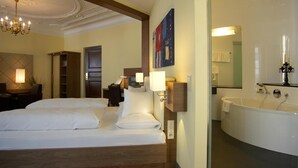 Ein Hotelzimmer mit einem großen Bett, einer Sitzecke mit Stuhl und Tisch, einem Bild an der Wand und einem Badezimmer mit Badewanne und Spiegel.