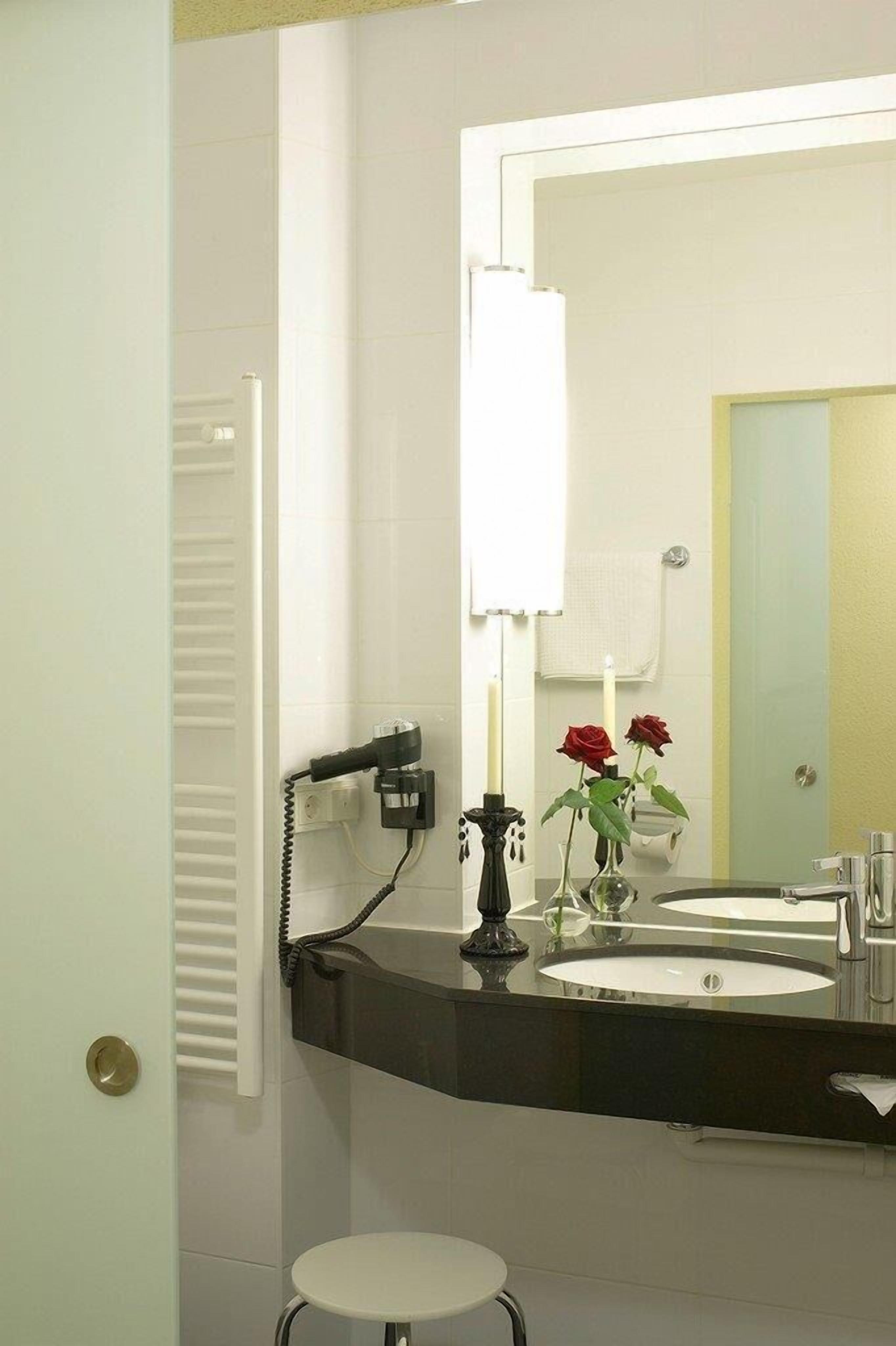 A bathroom with a white tiled wall, a black countertop, a round sink, a mirror, a towel rack, and a small white stool.