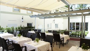 Ein Essbereich im Freien mit Tischen für zwei Personen, der von einem Baldachin überspannt und mit Stühlen umgeben ist.