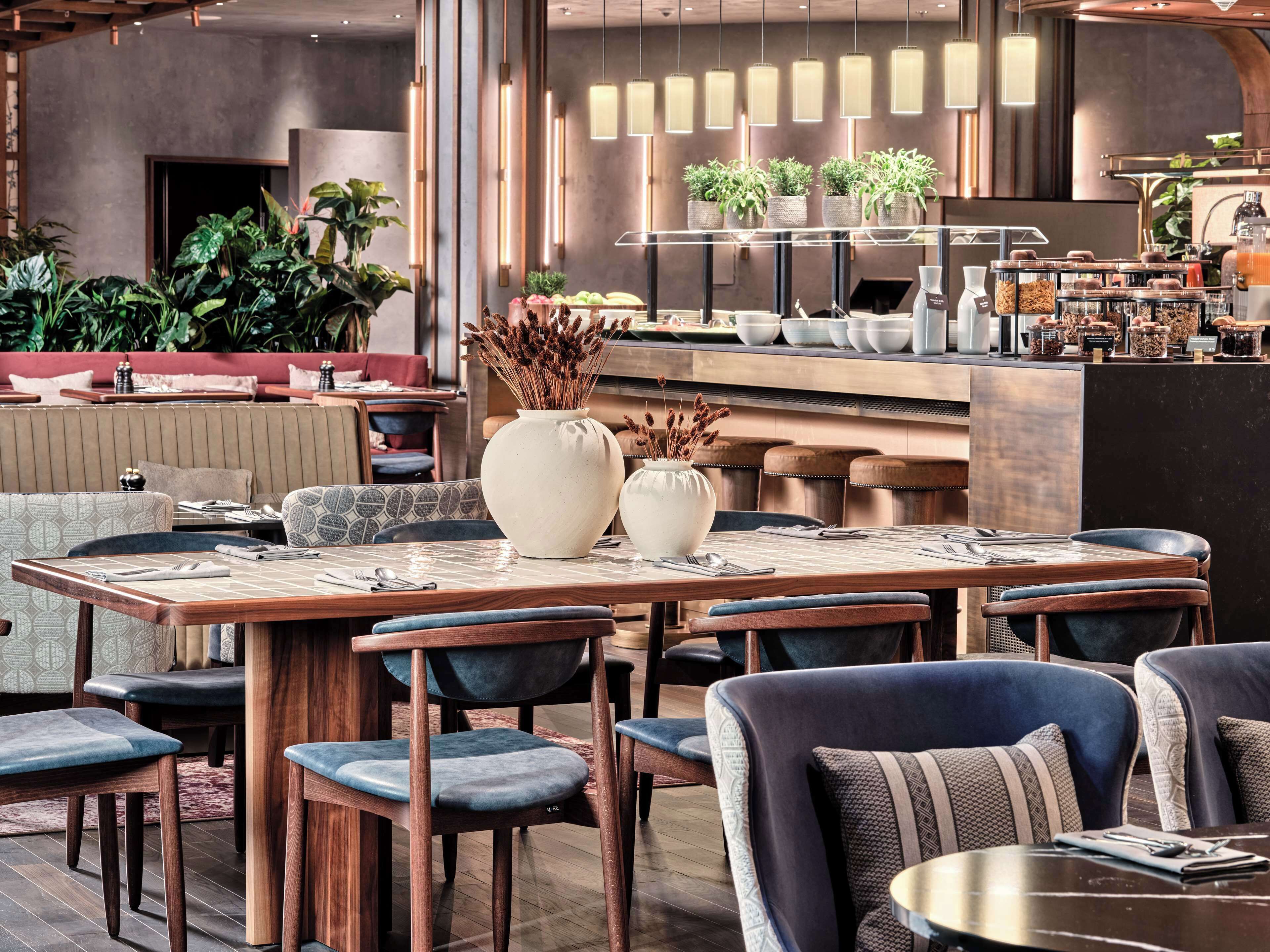 A modern restaurant with wooden tables, blue upholstered chairs, and a bar area with plants and food containers.