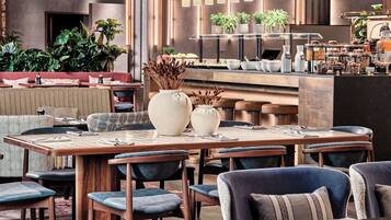 A modern restaurant with wooden tables, blue upholstered chairs, and a bar area with plants and food containers.