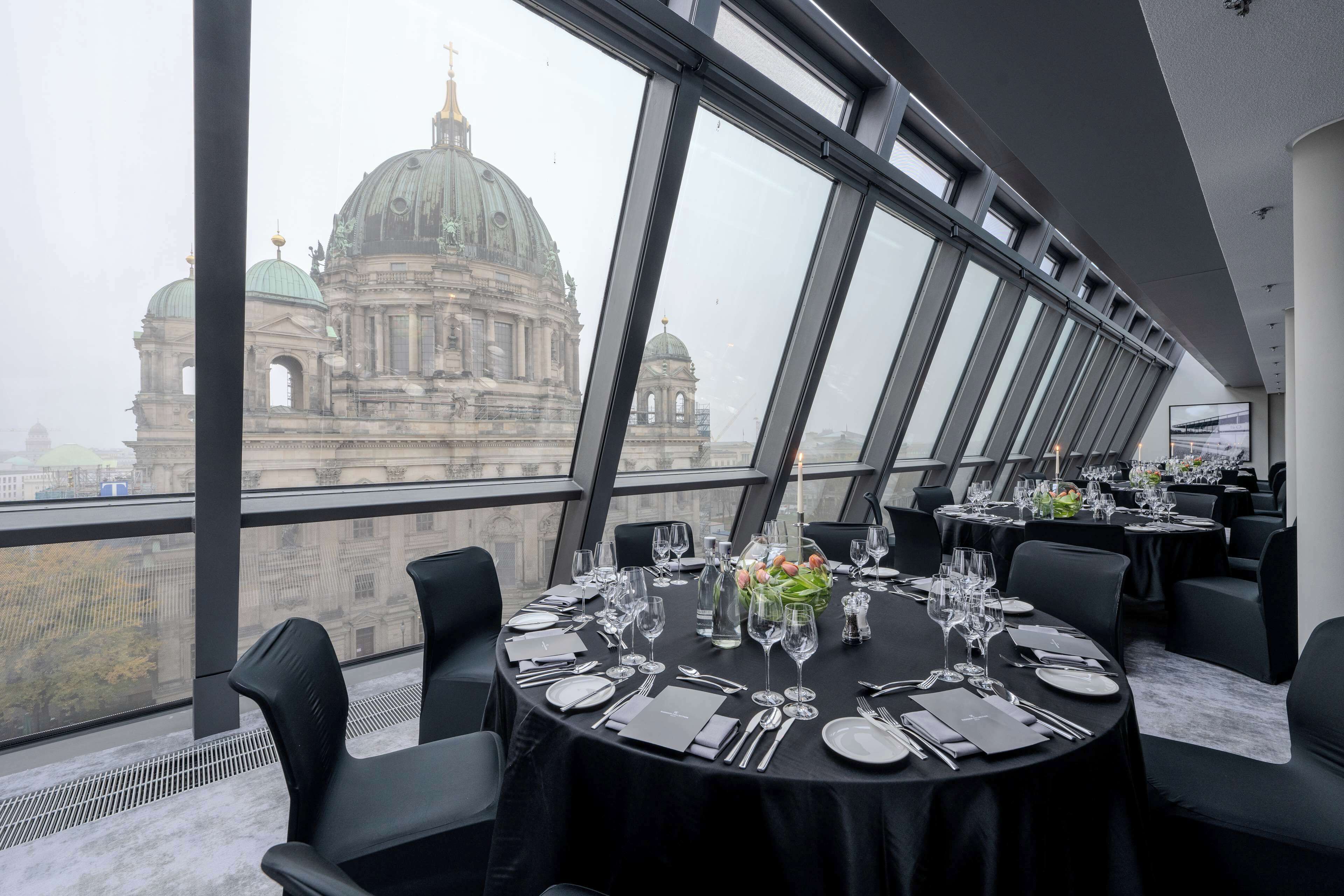 Un comedor con mesas redondas dispuestas para una comida, con ventanas amplias que ofrecen vistas a un edificio histórico.