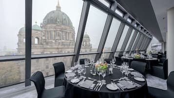 Un comedor con mesas redondas dispuestas para una comida, con ventanas amplias que ofrecen vistas a un edificio histórico.