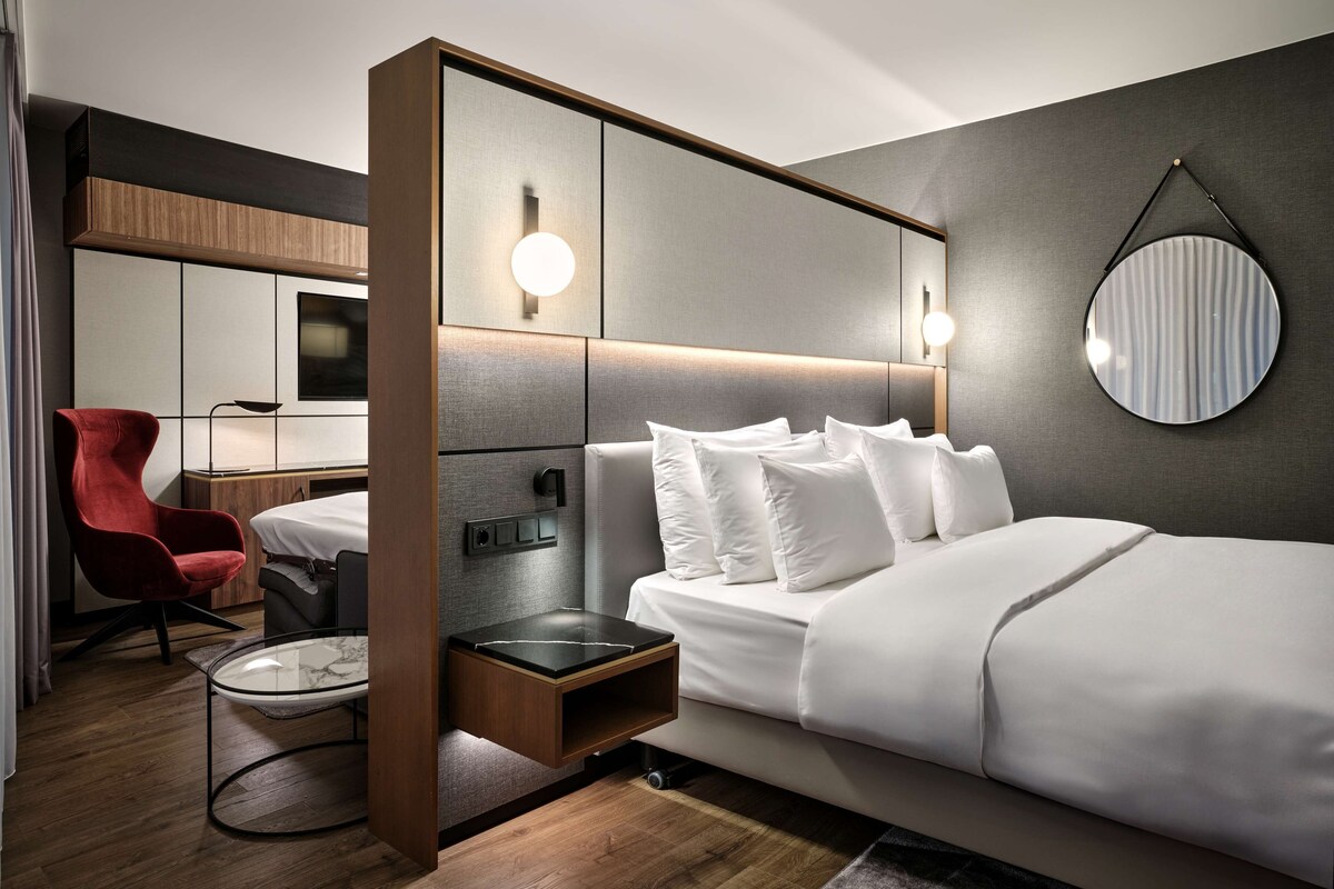 Een moderne hotelkamer met een groot bed, een bureau met stoel, een rode fauteuil en een ronde spiegel aan de muur.