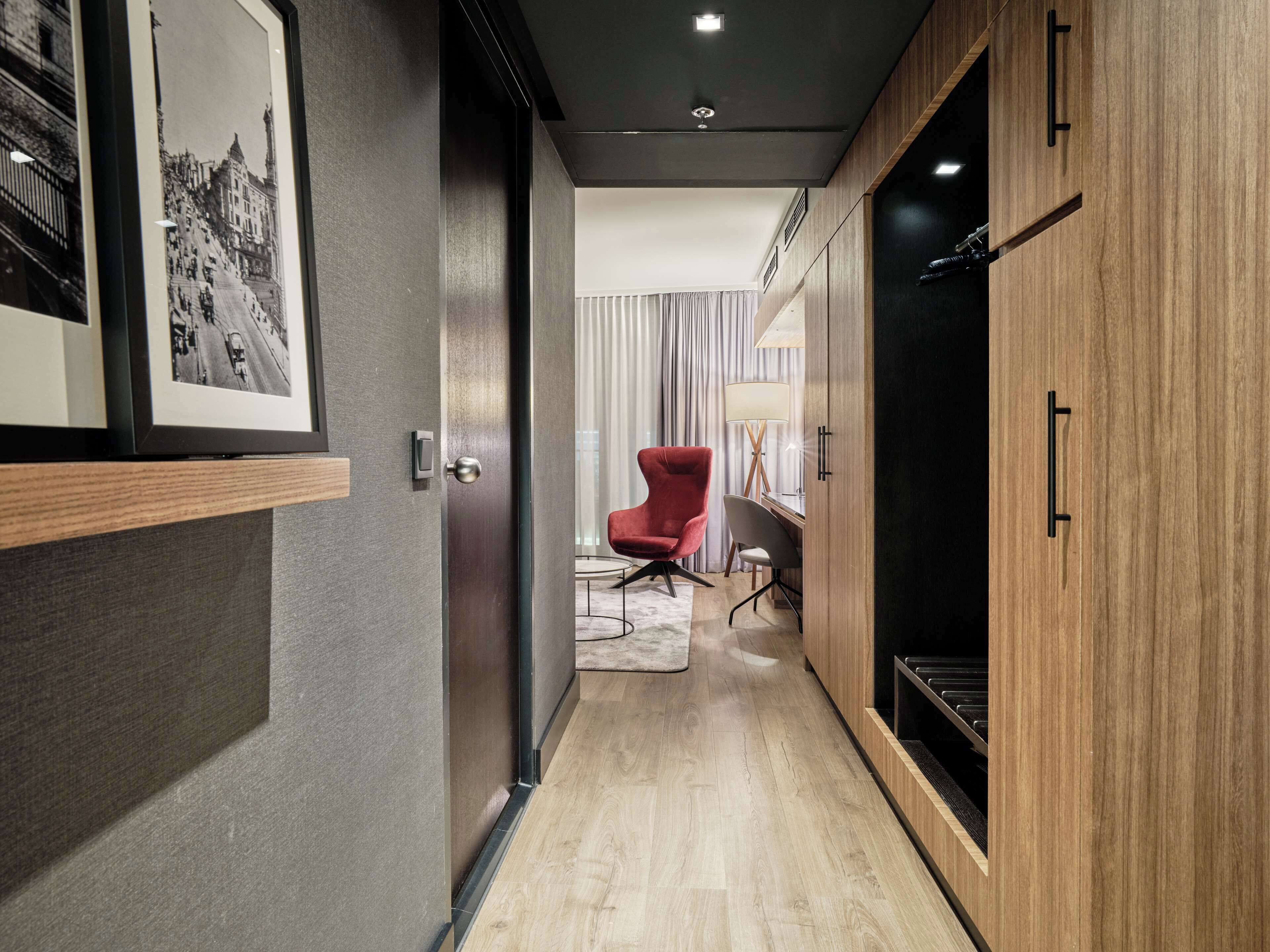 A modern hotel hallway with a wooden wardrobe, a red chair, and a desk with a lamp.